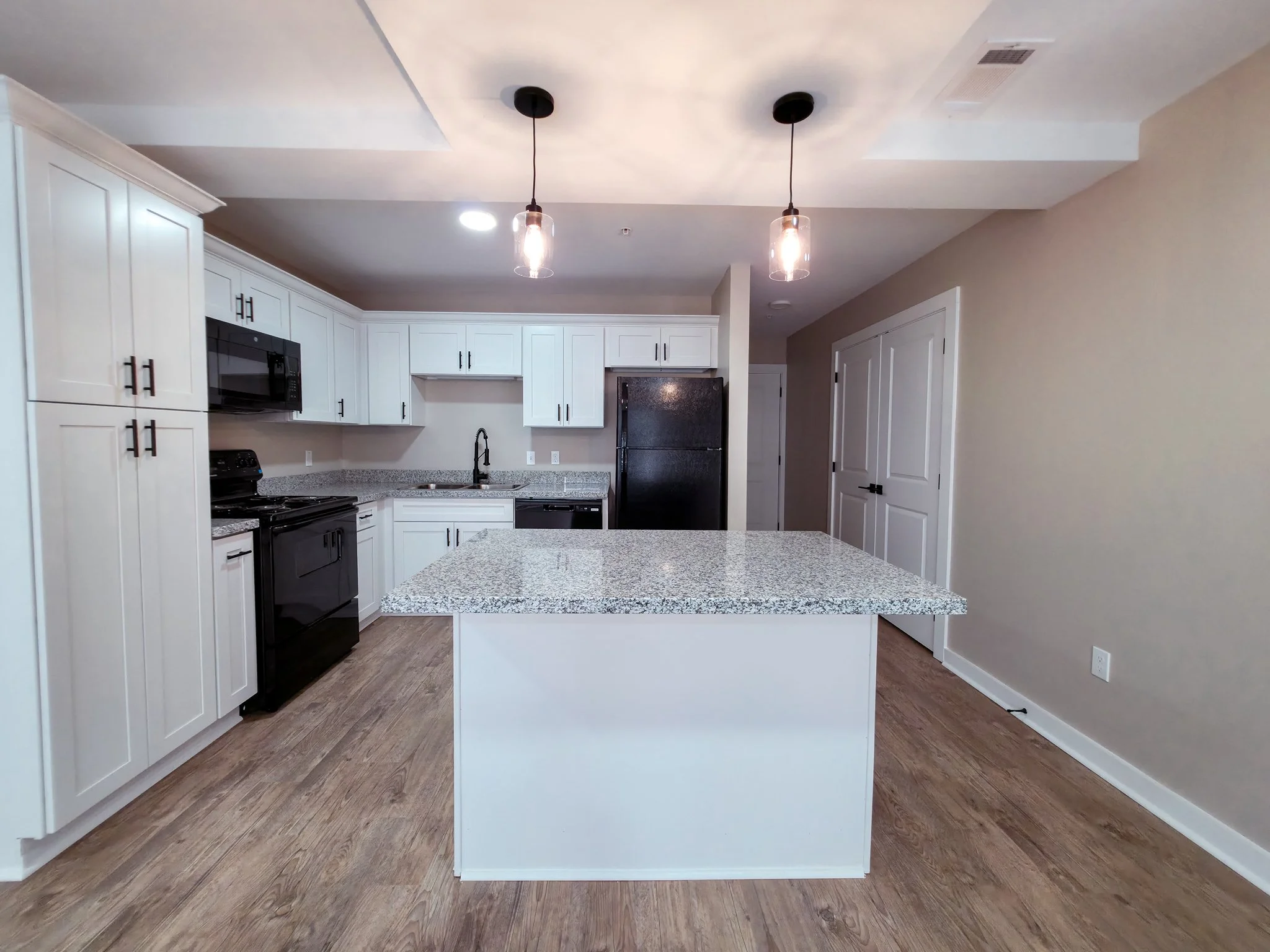 Modern kitchen with white cabinets, black appliances, granite countertops, and a central island, with pendant lighting and wood flooring.