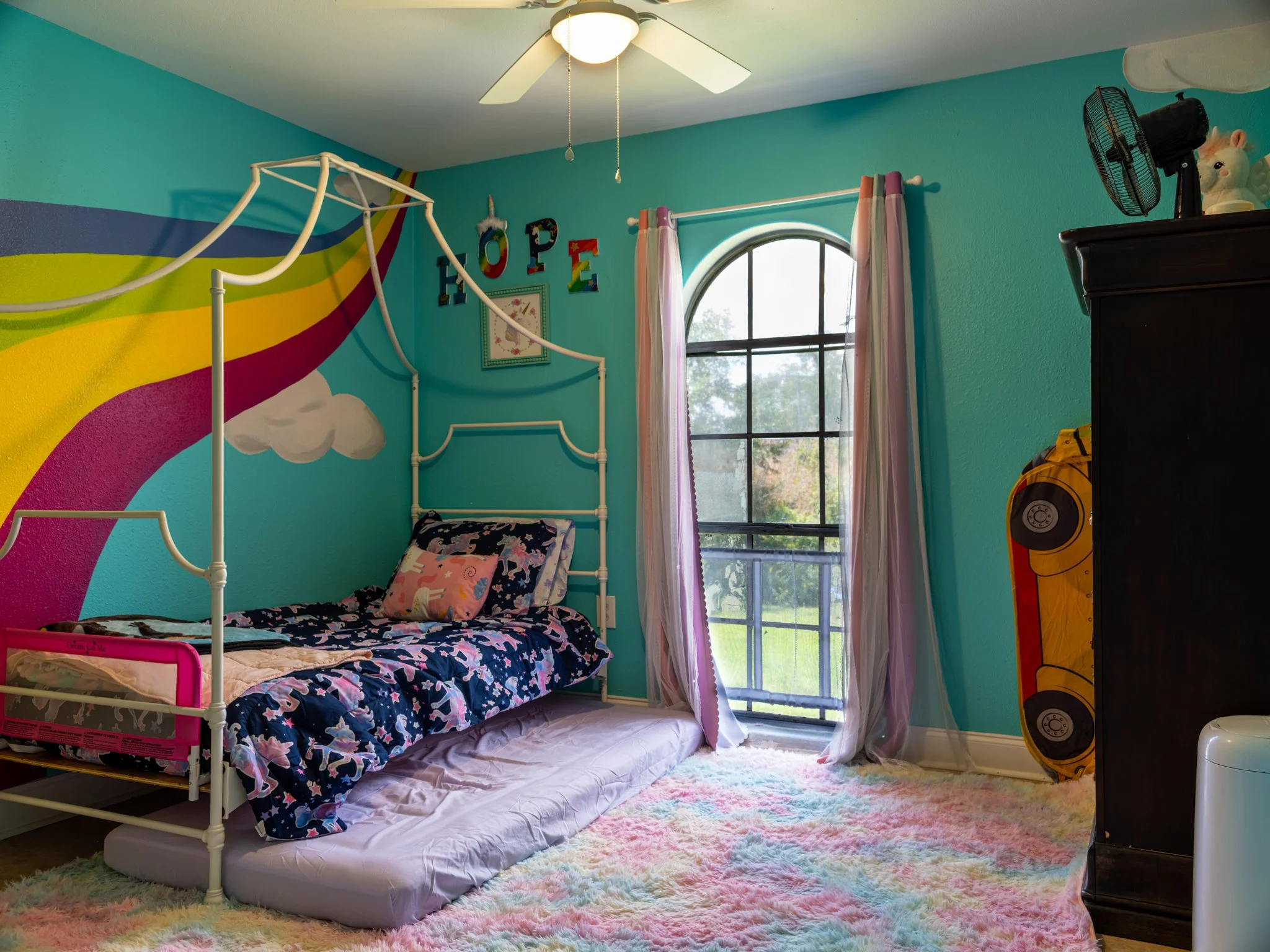 Colorful children's bedroom with rainbow mural on the wall, twin bed with navy unicorn bedding, pink unicorn pillow, large window with pink and purple curtains, rainbow-colored shaggy rug, and black dresser with a fan and unicorn figurine on top.