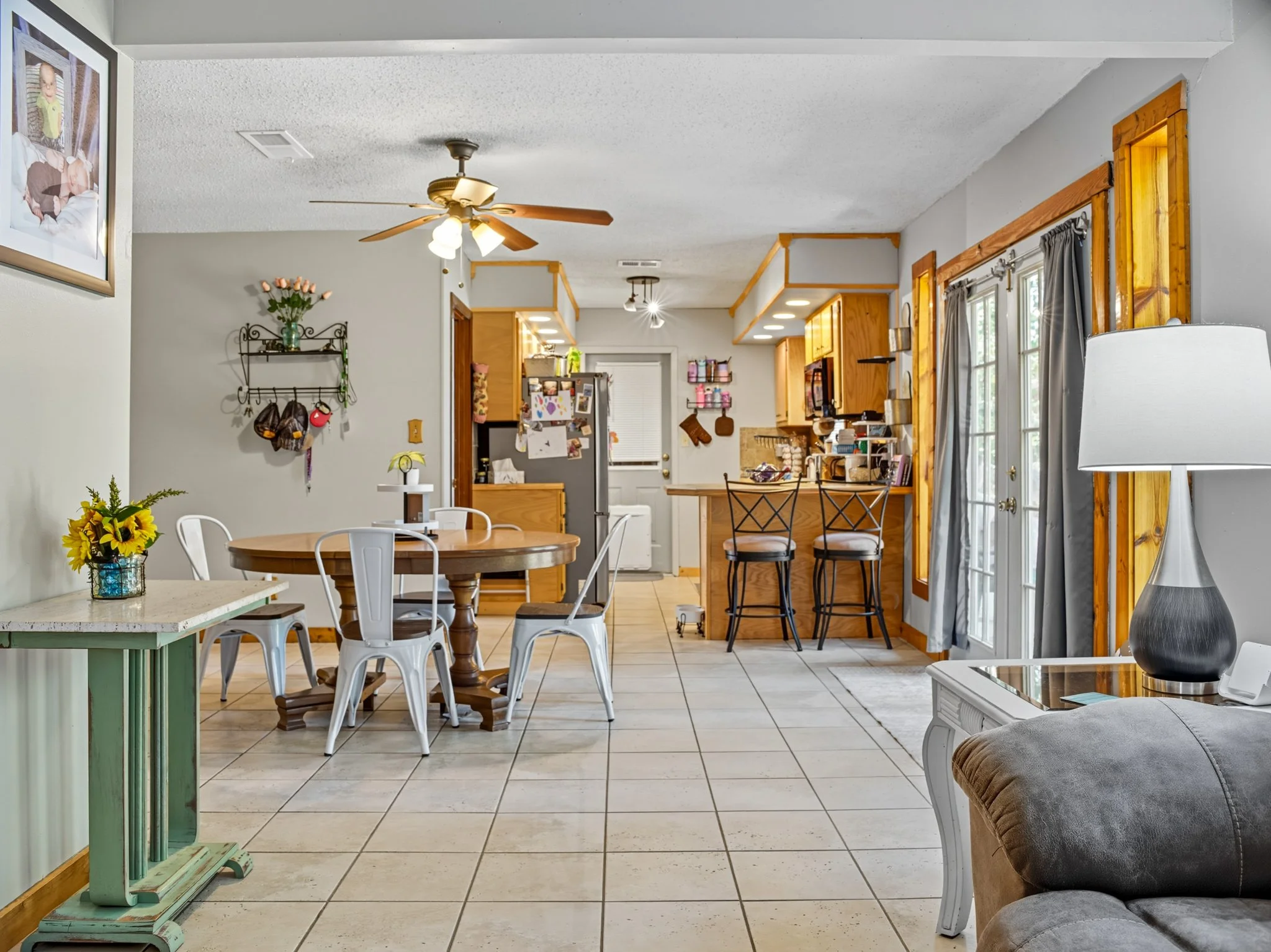 Open-concept kitchen and dining area with tiled floor, wood accents, bar stools, sliding glass door, and decorated with flowers and photos.