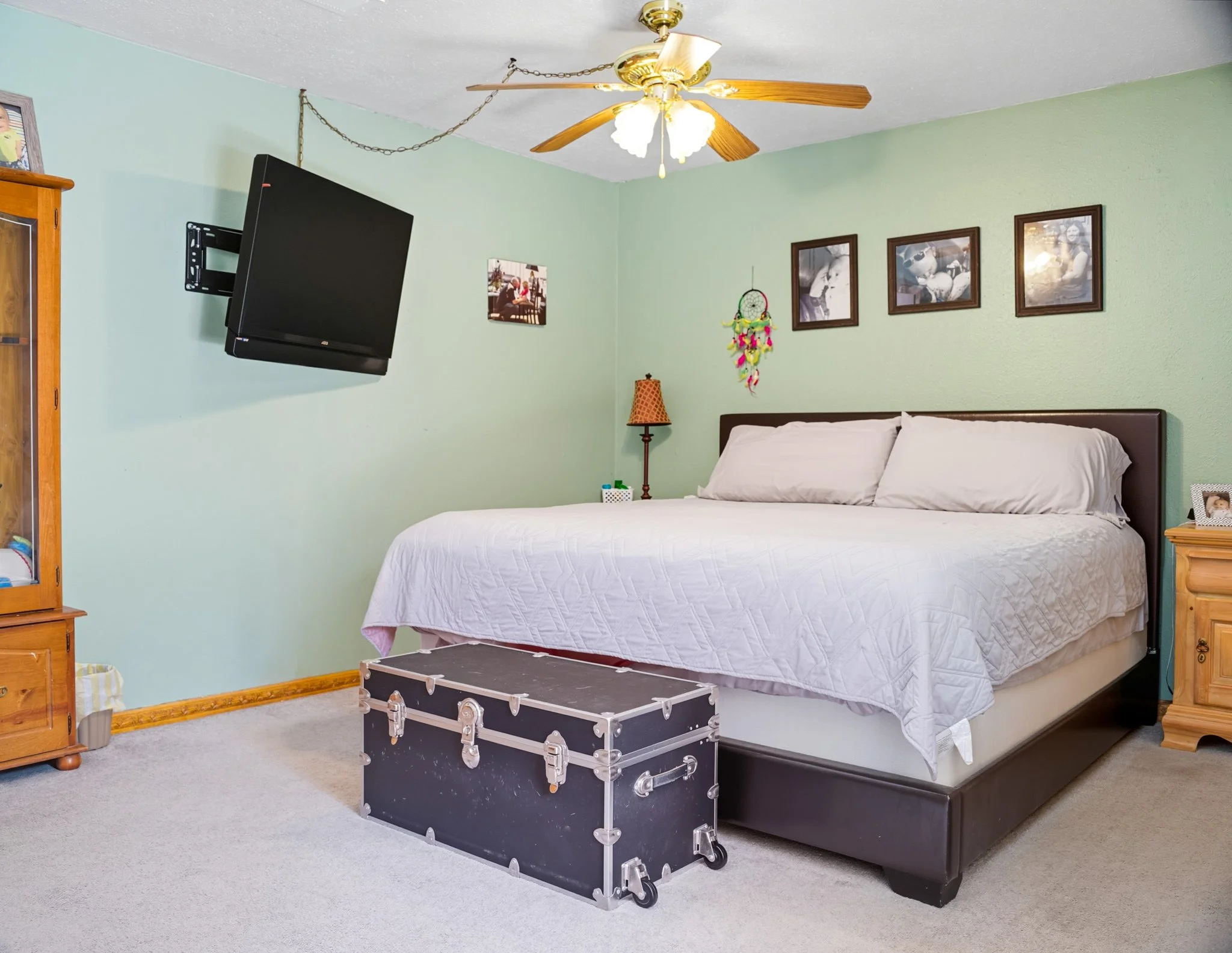 A bedroom with a green wall, a large bed with white bedding, a black storage trunk at the foot of the bed, a wall-mounted TV, a wooden cabinet, and pictures on the wall.