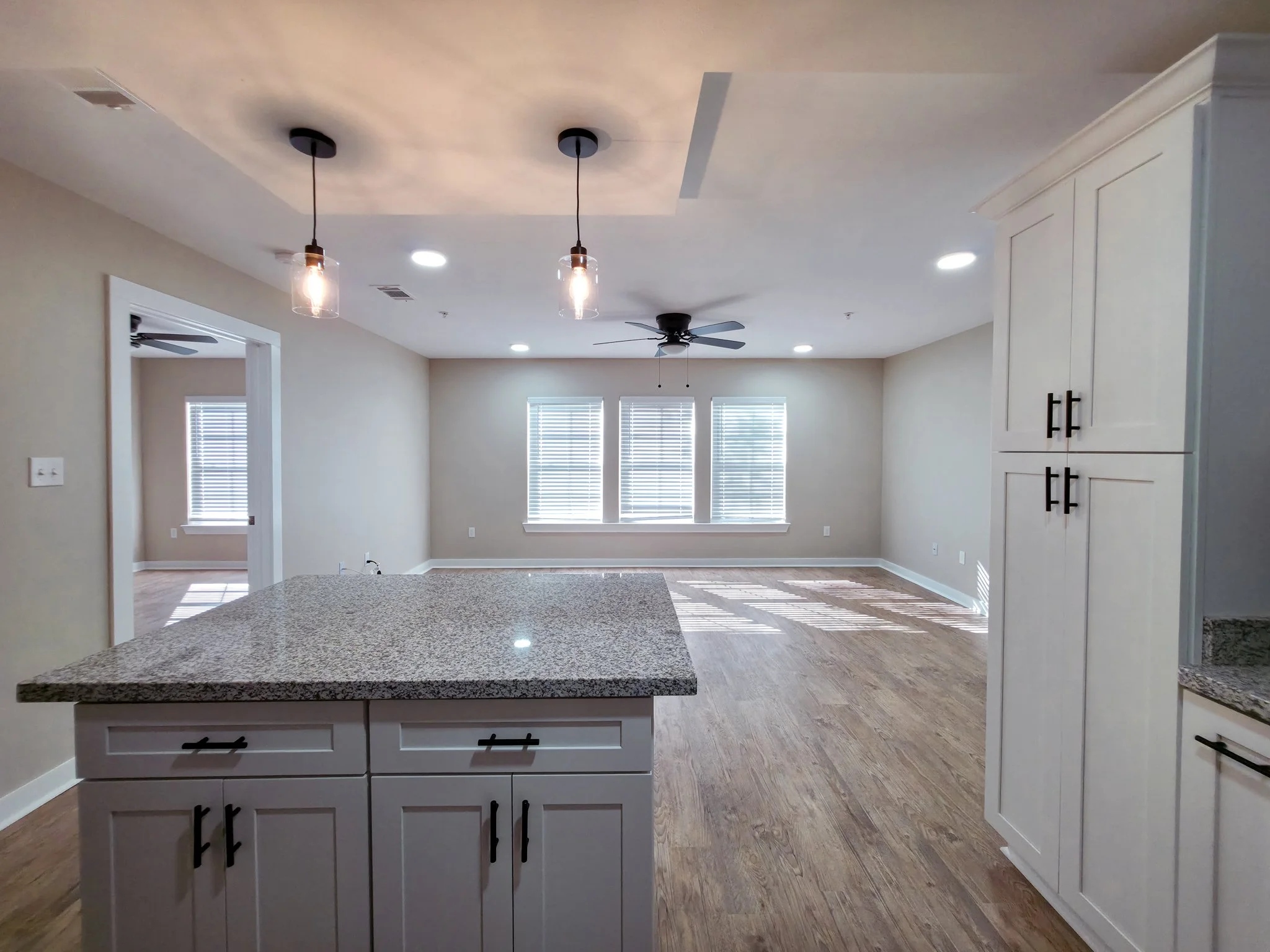 Empty living room with three large windows, ceiling fan, and hardwood floors. The view is from a kitchen area with a granite countertop and white cabinets.