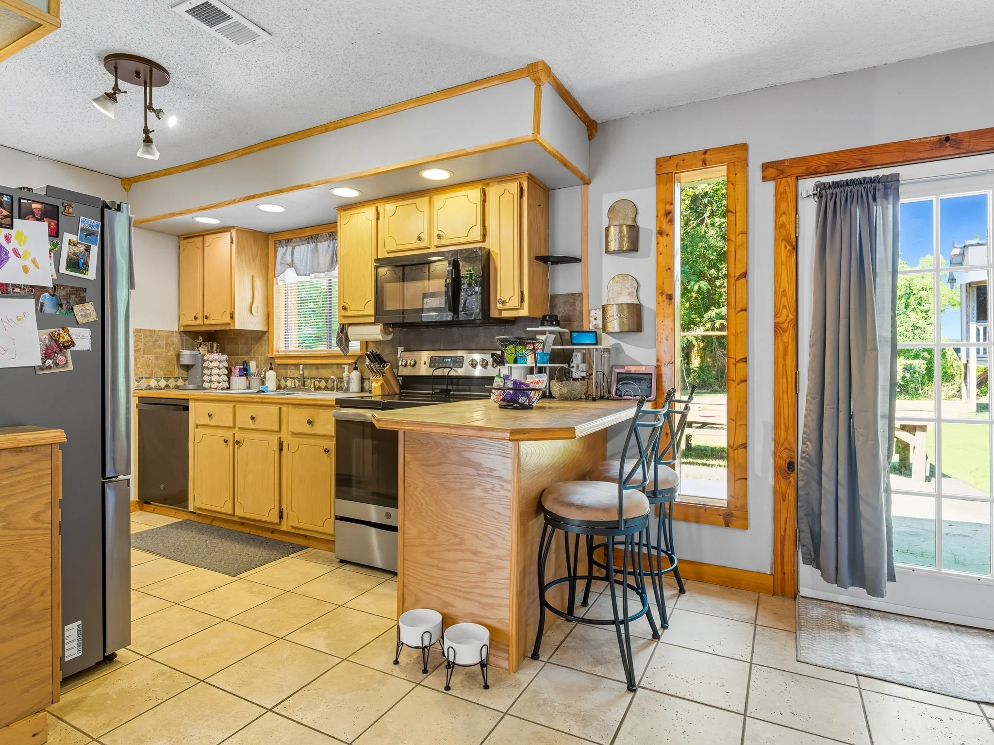 Kitchen with yellow cabinets, stainless steel stove and microwave, refrigerator with kids' drawings, wooden window frames, sliding glass door with curtain, tiled floor, breakfast bar with bar stools, and outdoor view with greenery.