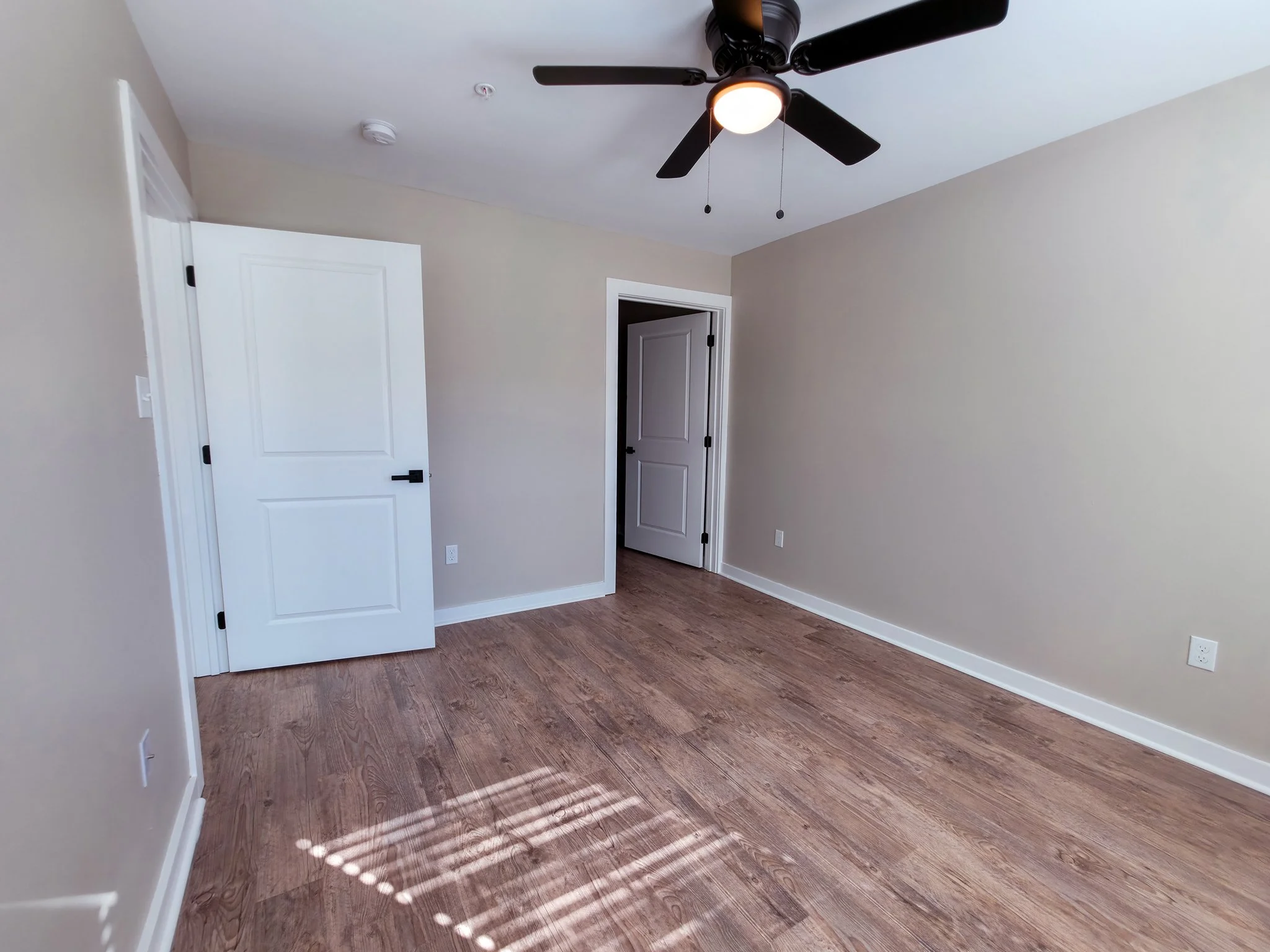 Empty room with beige walls, hardwood floors, an open white door, a ceiling fan with light, and sunlight streaming through window blinds.