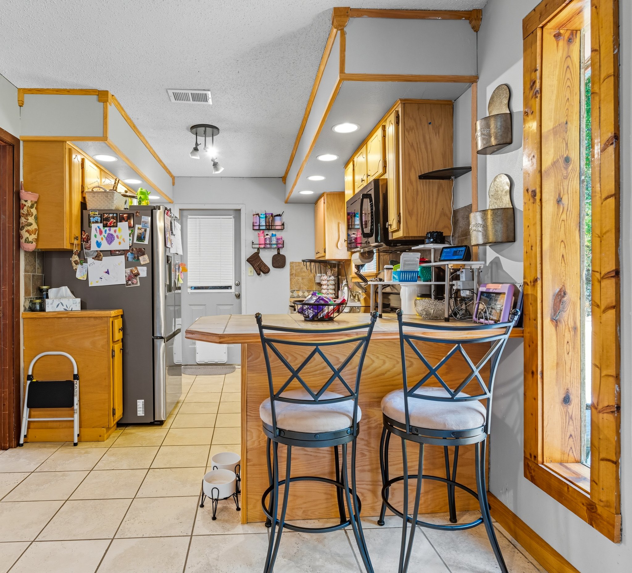 Kitchen with wooden cabinets, stainless steel refrigerator covered with children's drawings, a small bar with two barstools, and various kitchen items on the counter. A door with a window and a small folding step stool to the left of the refrigerator