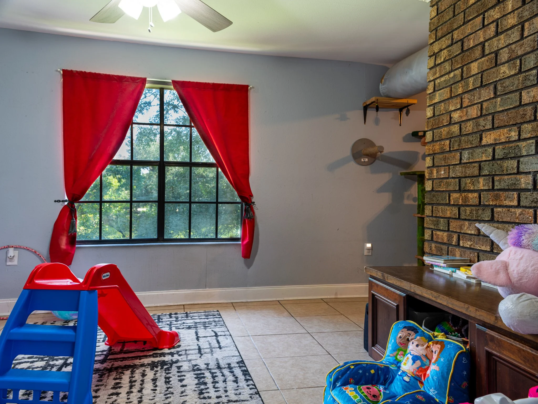 A children's playroom with a window featuring red curtains, a red and blue plastic slide, a colorful rug, a brick accent wall, a wooden cabinet with toys, and a plush unicorn stuffed animal.