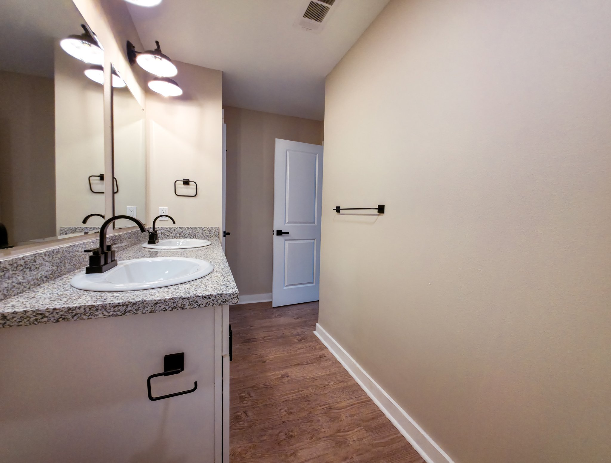 Bathroom vanity with granite countertop, two sinks, and black fixtures, beige walls, wood-look flooring, towel rack on wall, open door in background.