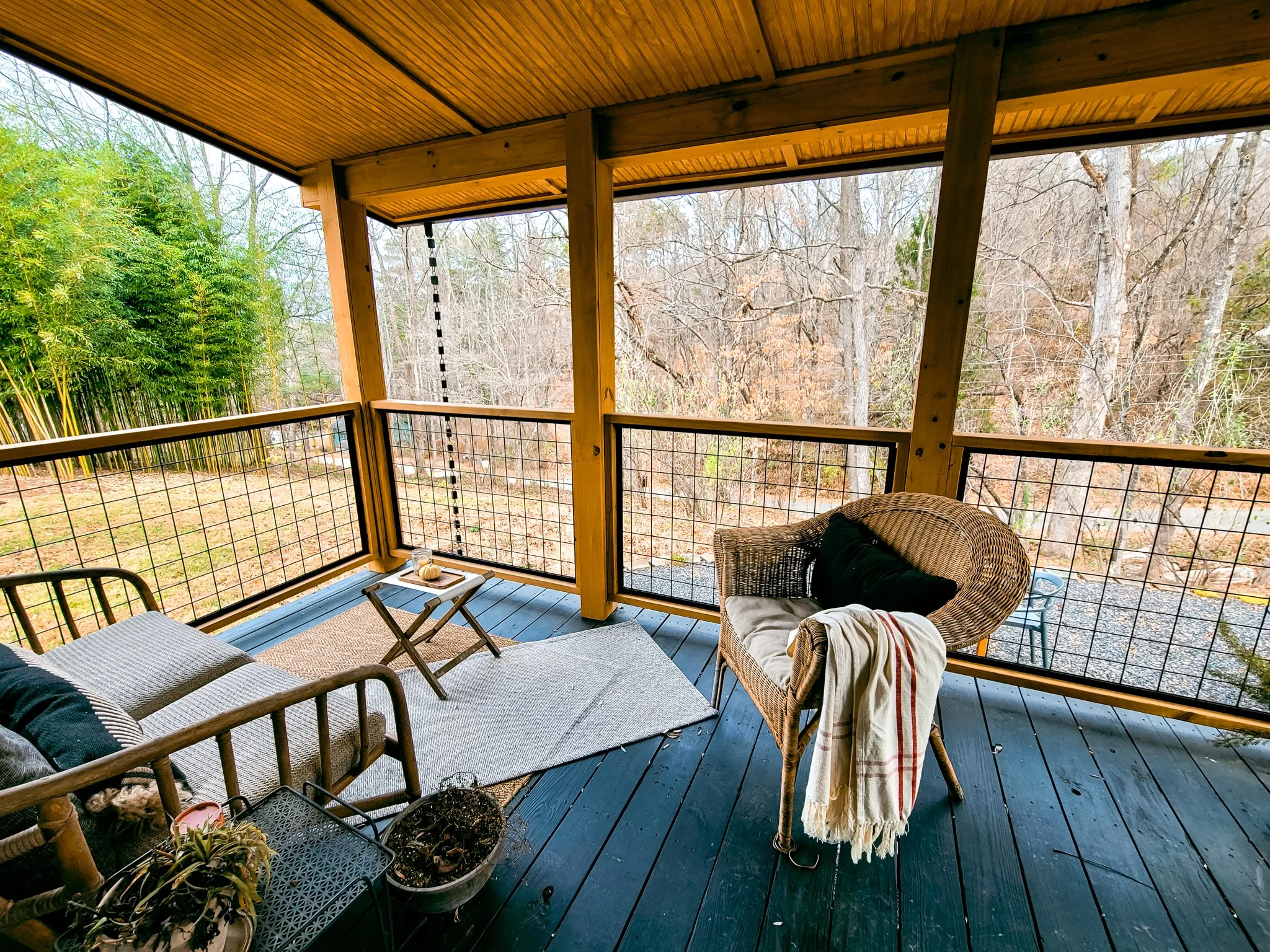 Covered porch with outdoor seating including a wicker chair with blanket, a wooden bench, and a small table, overlooking a wooded area with trees.
