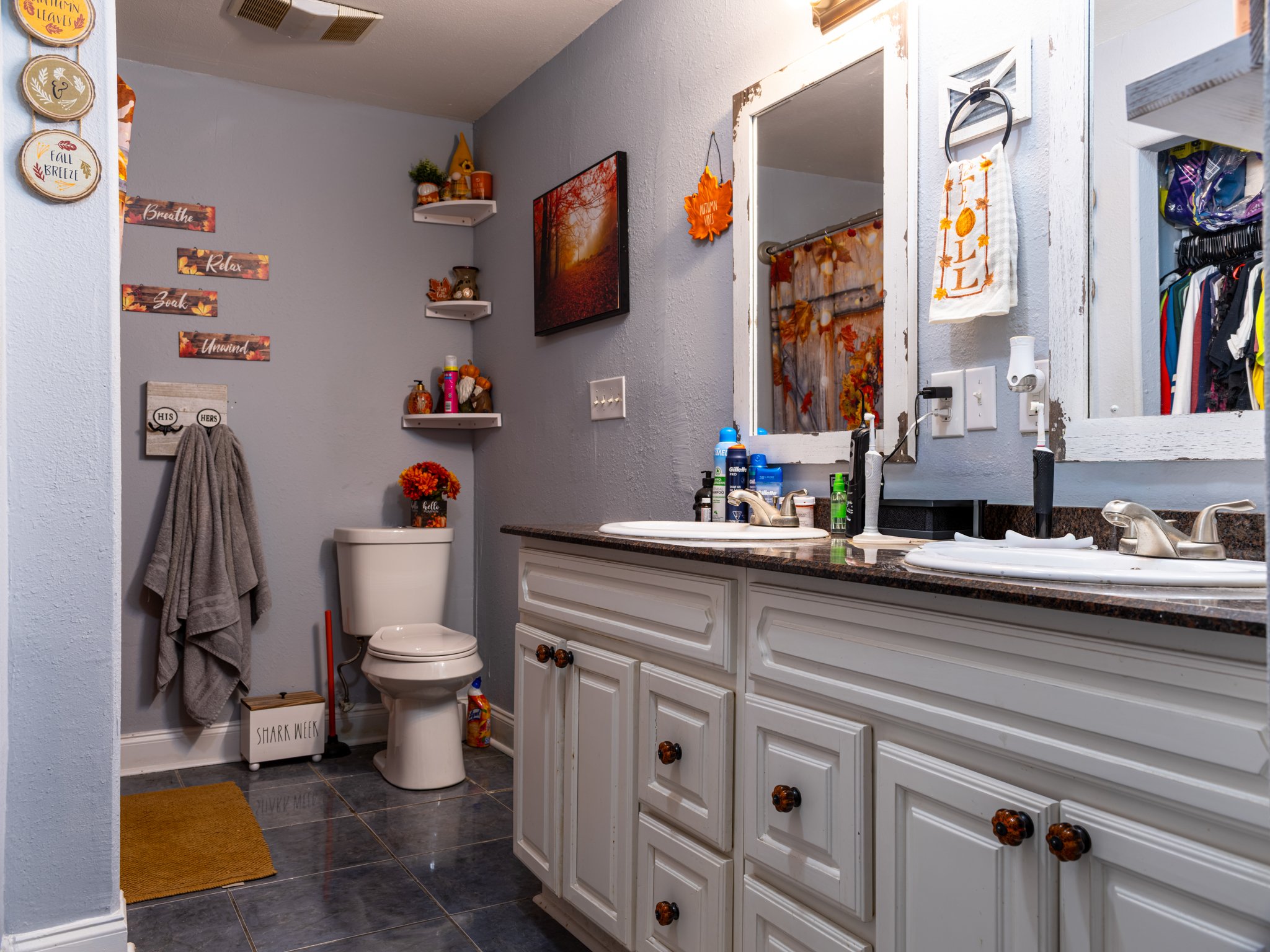Bathroom with double sink vanity, large mirror, gray walls decorated with fall themed decorations, toilet with fall flowers, enclosed closet with clothing, and wall shelves with seasonal decor.