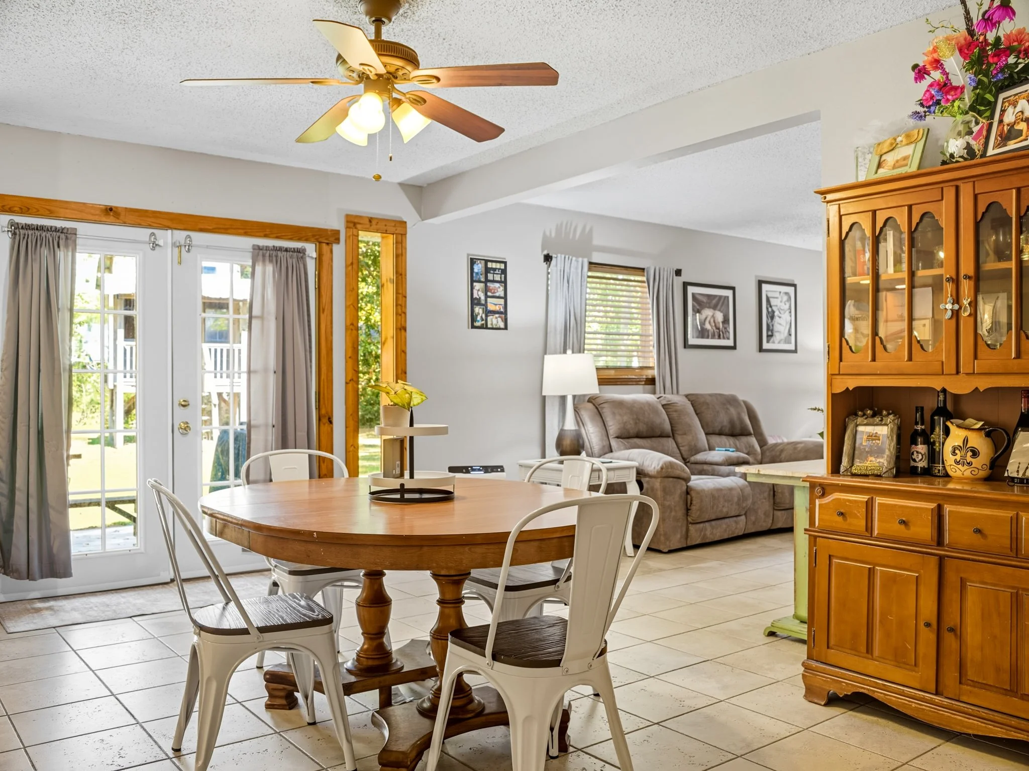 A dining area with a round wooden table and four white chairs, adjacent to a living room with a sectional sofa, a lamp, and framed pictures on the wall. There's a ceiling fan with lights, a wooden china cabinet with decorations, and glass doors leadi