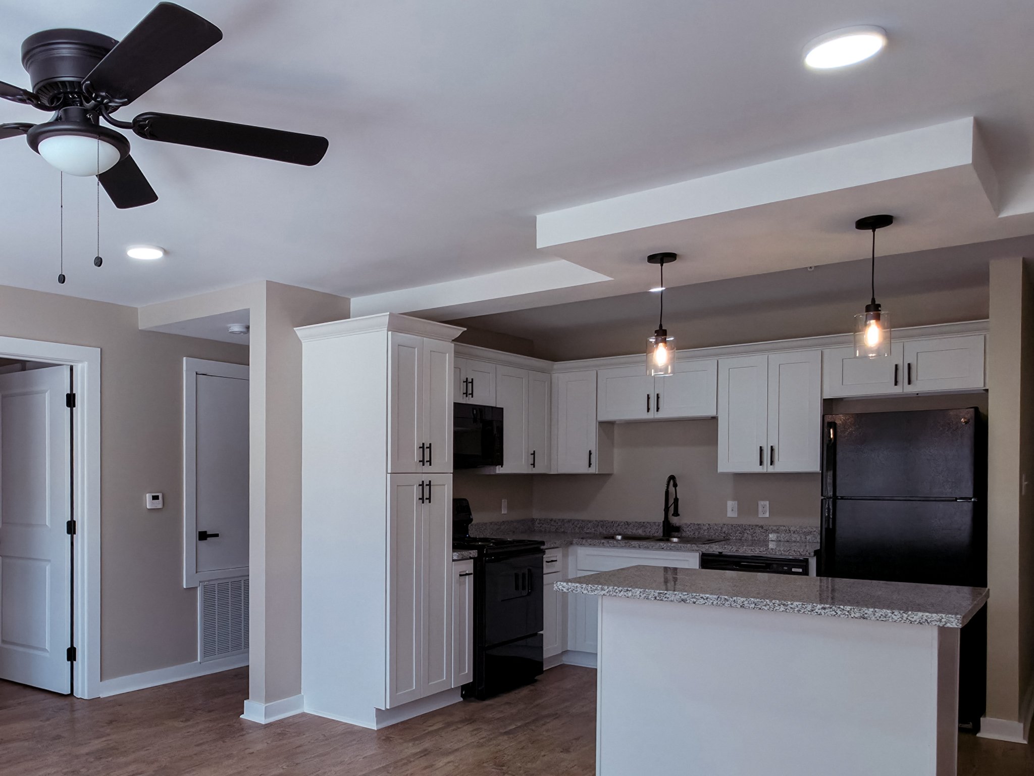 Modern kitchen with white cabinets, granite countertops, black appliances, pendant lights, ceiling fan, and hardwood floors.