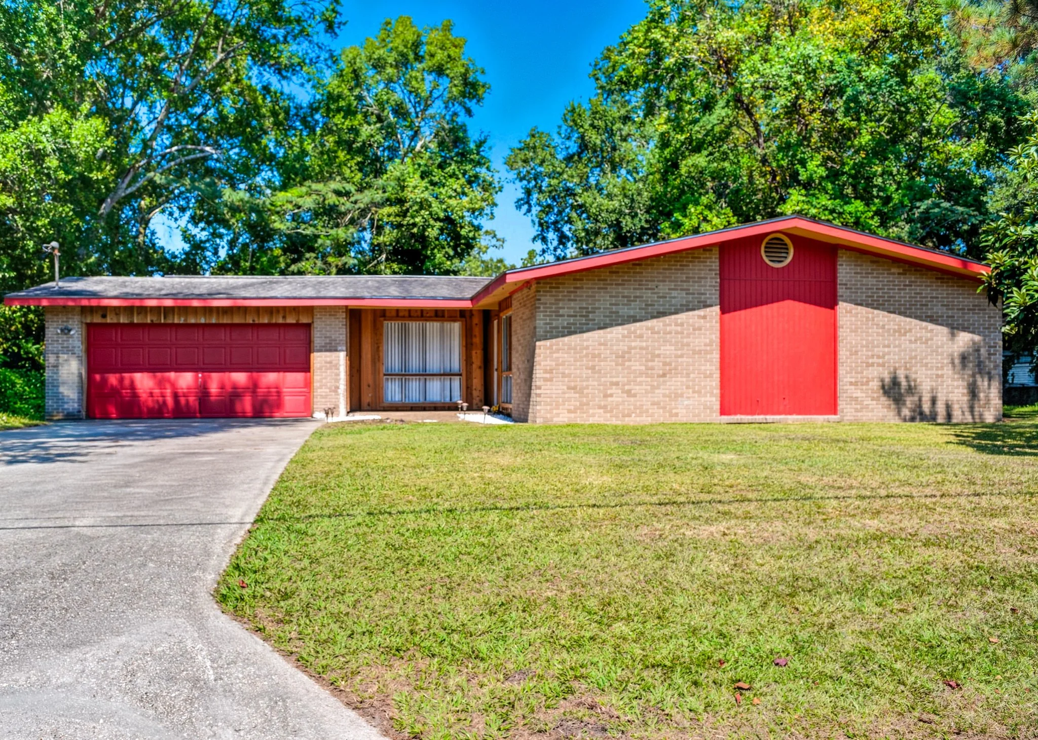 A single-story house with a red garage door, a red sliding door, and red accents on the roof and wall. The house has a concrete driveway and a well-maintained lawn, with tall green trees in the background and blue sky overhead.
