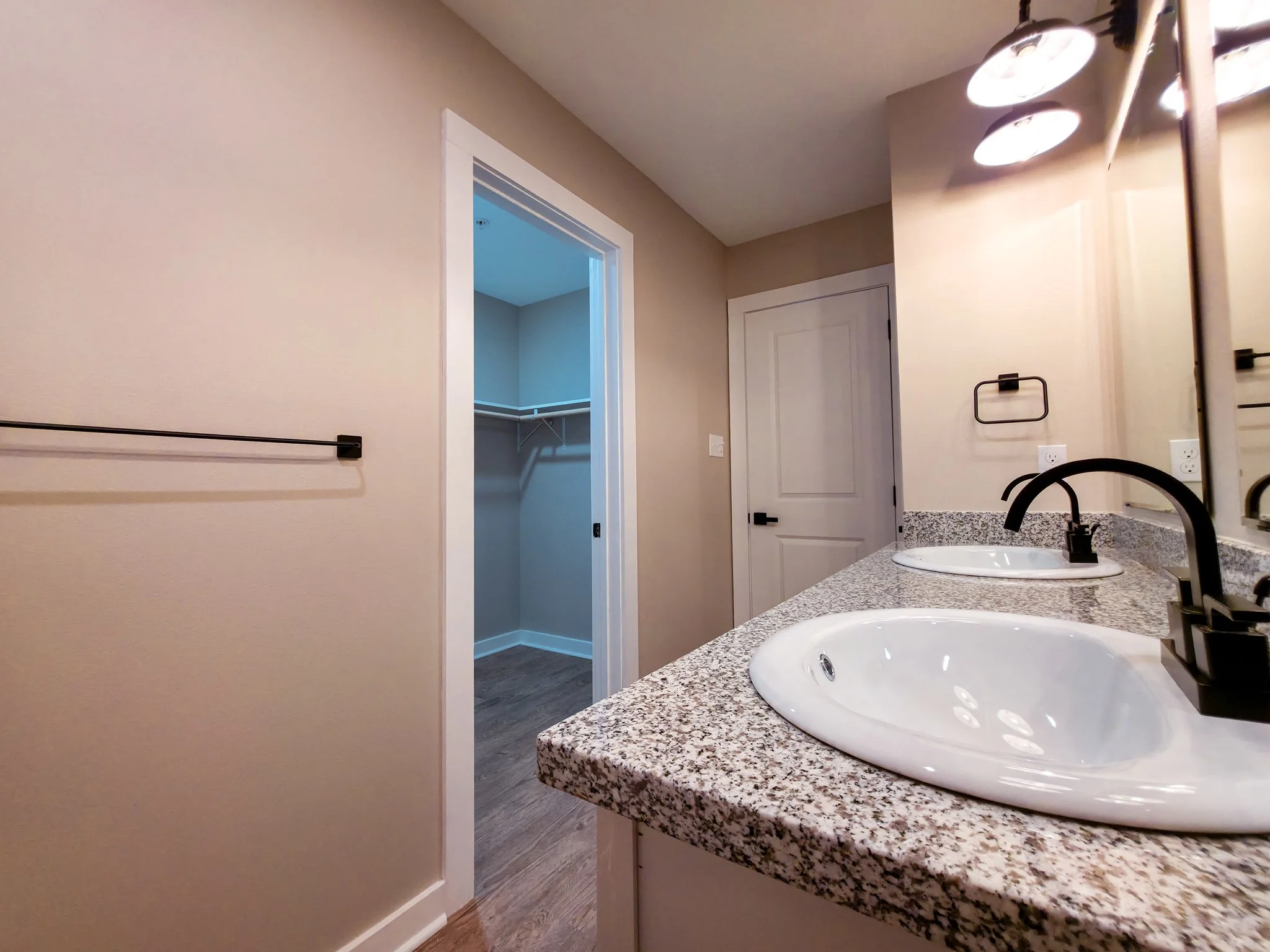Bathroom with granite countertop, two white sinks, black faucets, large mirror, beige walls, and an open walk-in closet with a closet rod inside.
