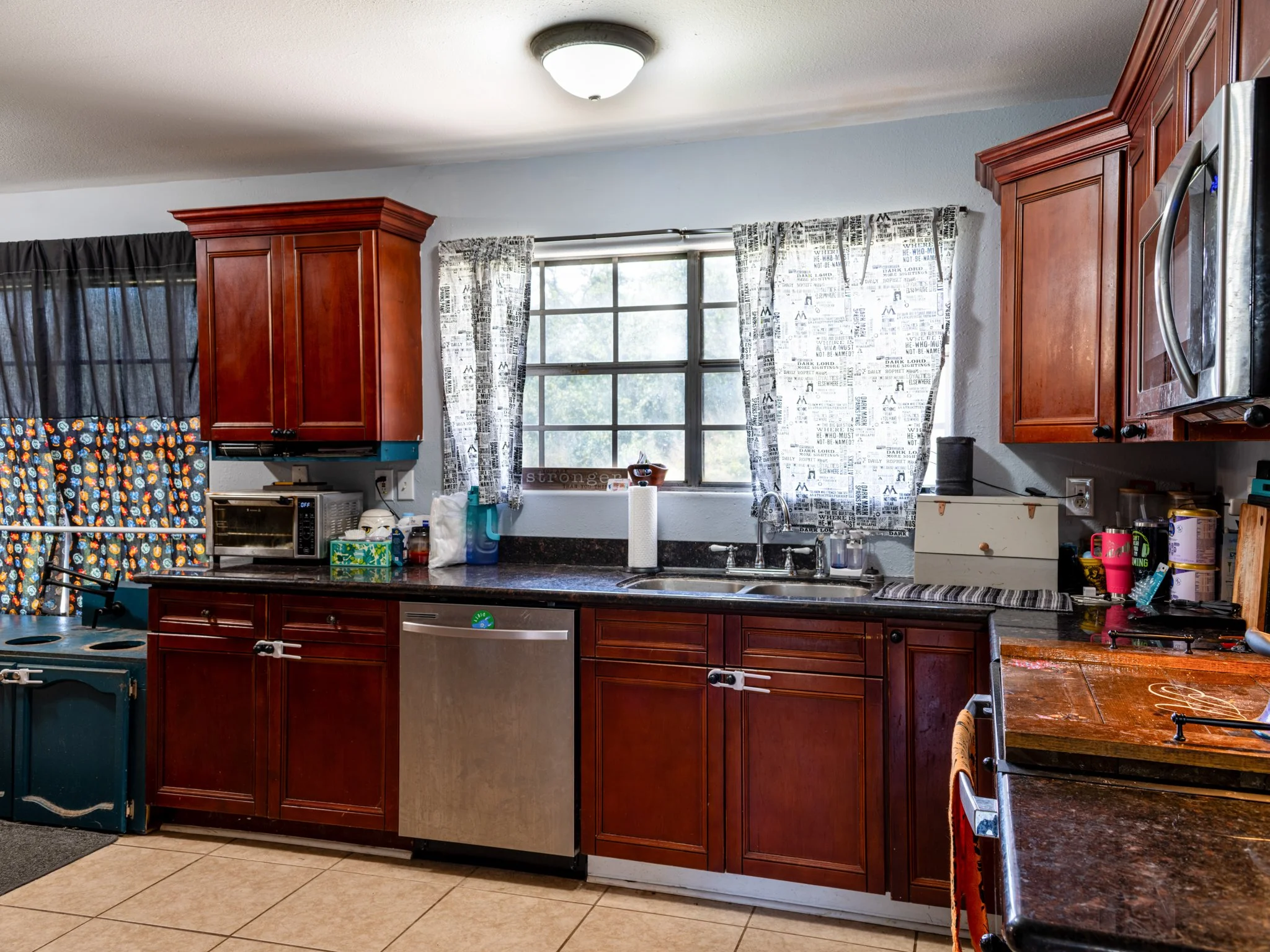 A kitchen with dark wood cabinets, a window with white and black patterned curtains, a granite countertop, a double sink, and various appliances and items on the counter.