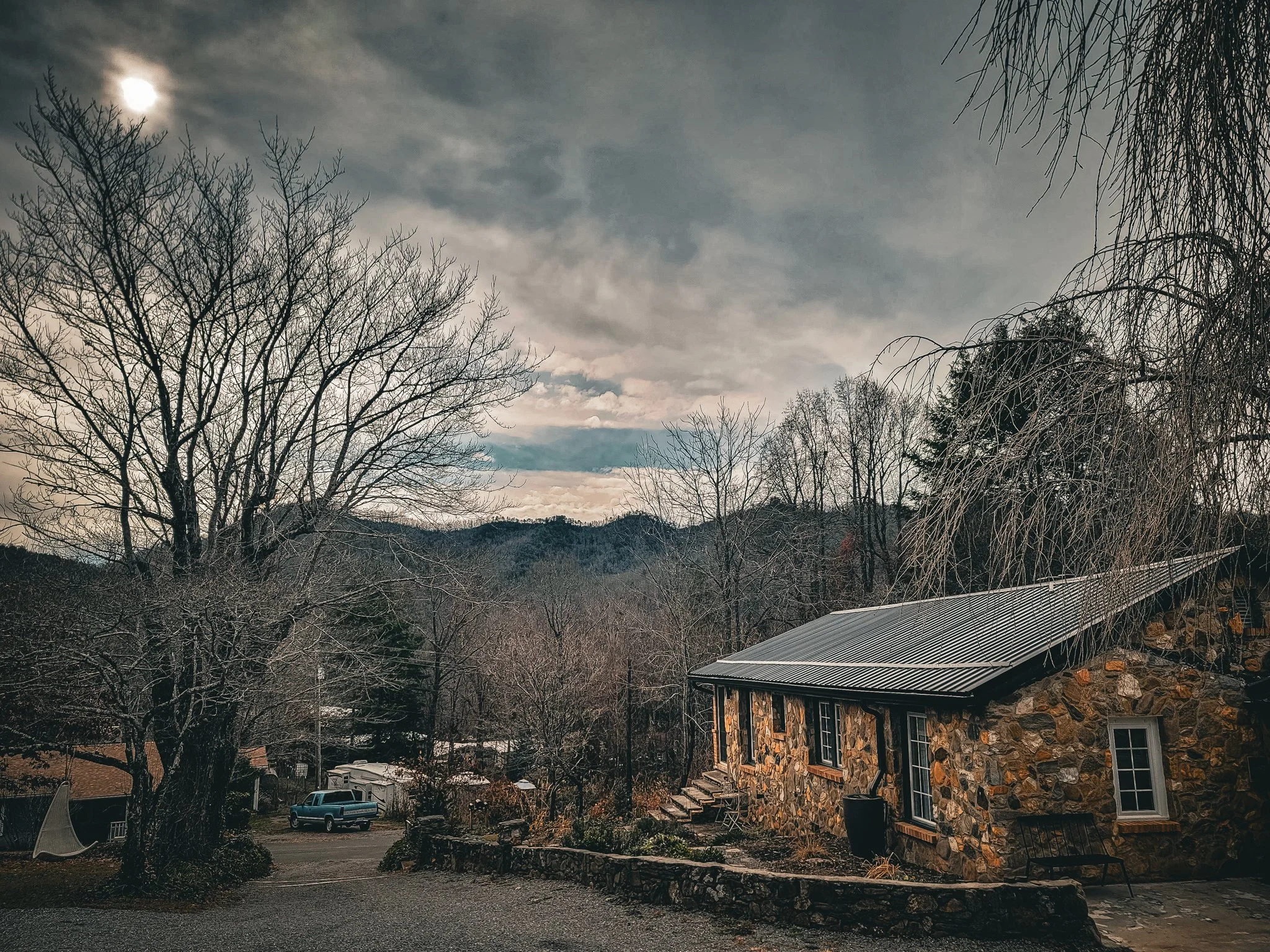 A rural scene with leafless trees, a stone house with a metal roof, a parked truck, and a backdrop of hills under a cloudy sky with the moon visible.