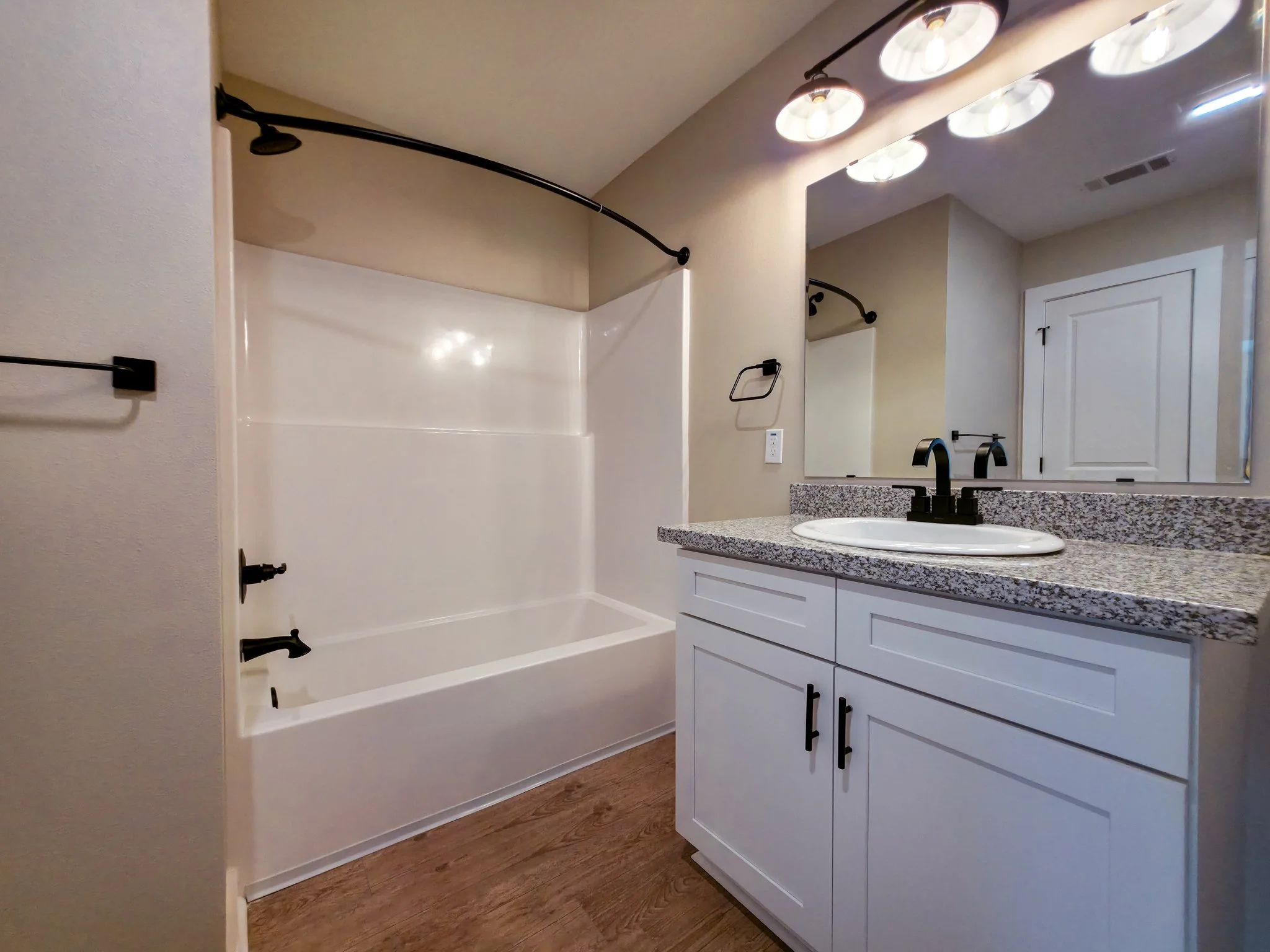 Bathroom with white vanity, granite countertop, oval sink, black faucet, large mirror, overhead lights, and a bathtub with a curved shower curtain rod.