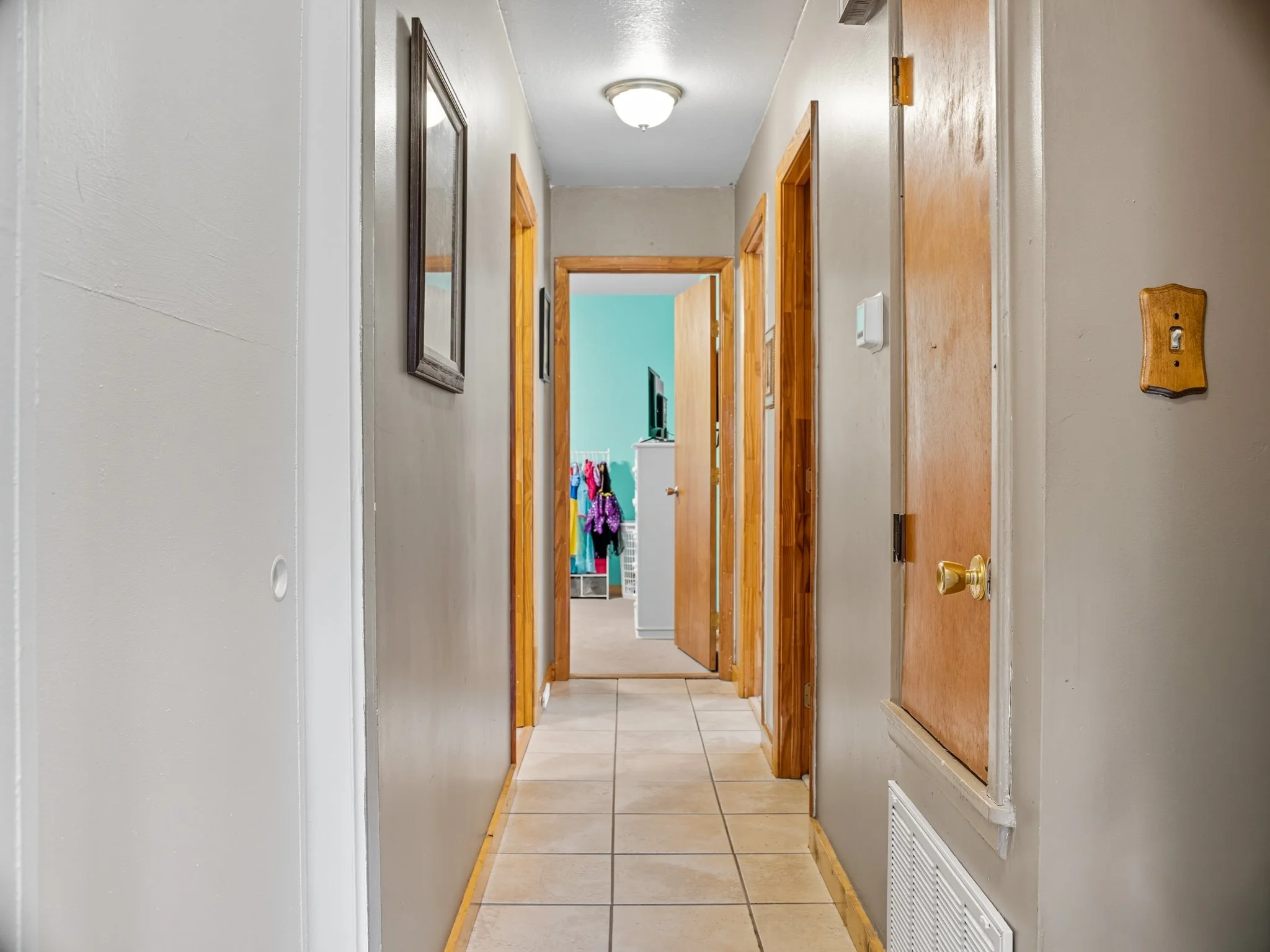A hallway in a house with beige tile flooring, light grey walls, wood trim around doors, and picture frames on the left wall. The end of the hallway opens into a room with a blue wall and a TV, with open doorways revealing a laundry basket and clothi
