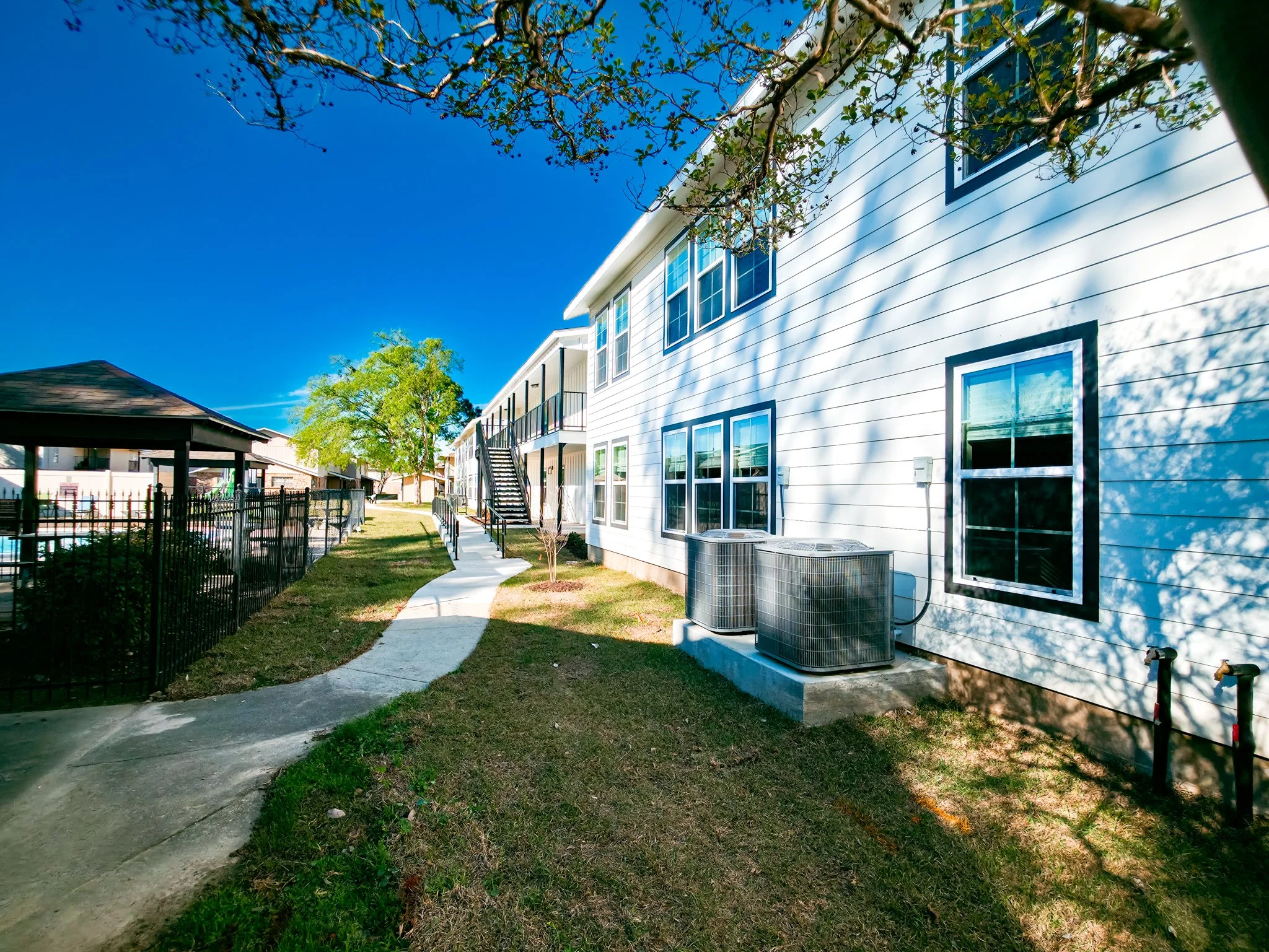 View of a multi-unit residential building with white siding, multiple windows, and air conditioning units outside. A sidewalk curves along the grassy yard, and there is a staircase leading to an upper level. The sky is clear and blue, and a tree cast