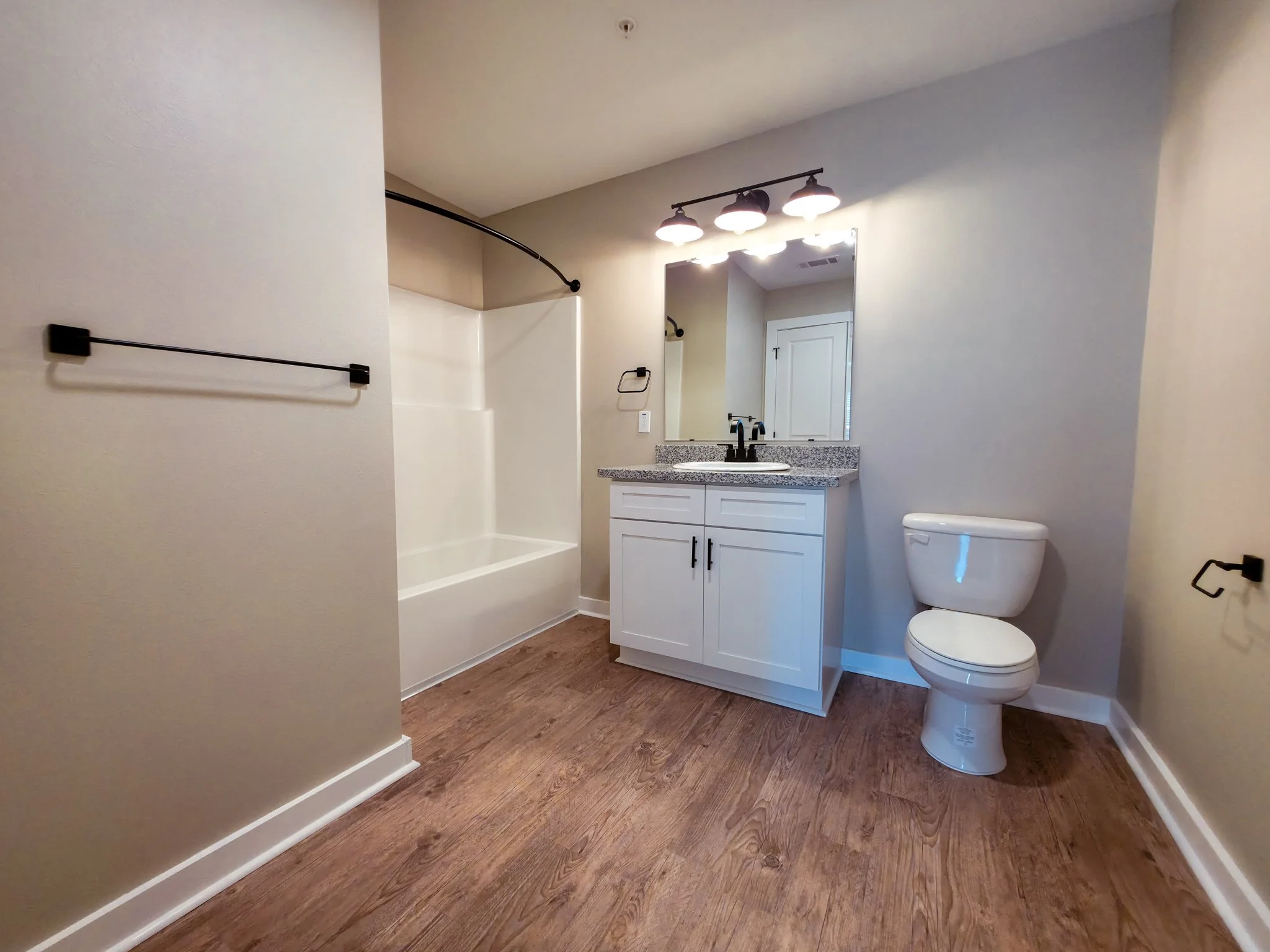 Bathroom with a bathtub and shower, a white vanity with a sink, a large mirror, a toilet, wood flooring, and wall-mounted towel racks.