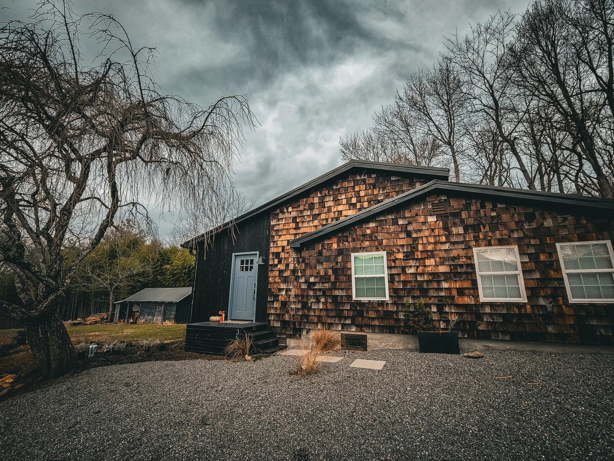 A rustic house with wood shingles and three white window panes, set against a dark, cloudy sky with leafless trees.
