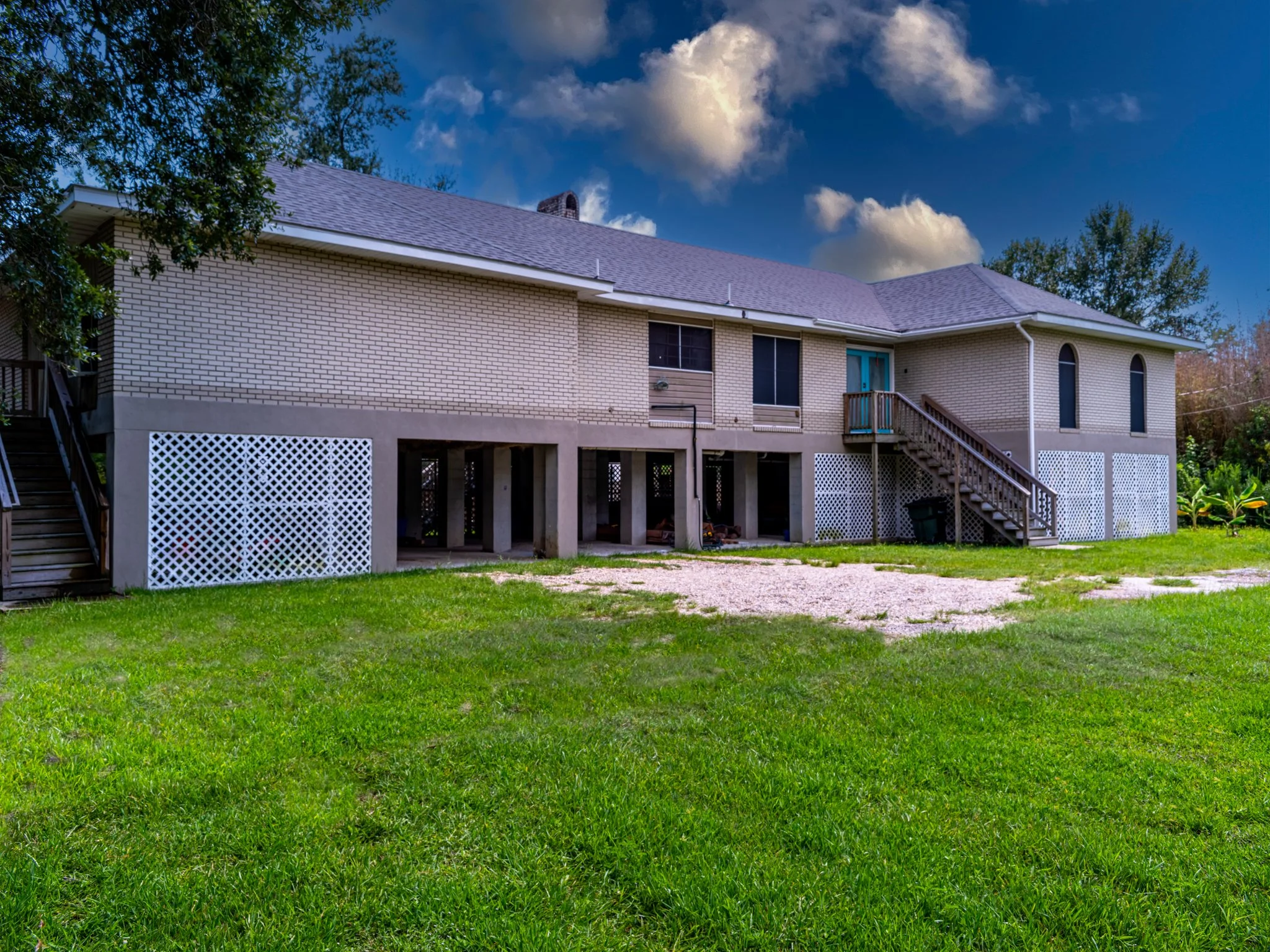 A two-story house with a brick exterior, elevated on concrete pillars, with stairs leading to a balcony door, green lawn, and tree on the left side under a partly cloudy blue sky.