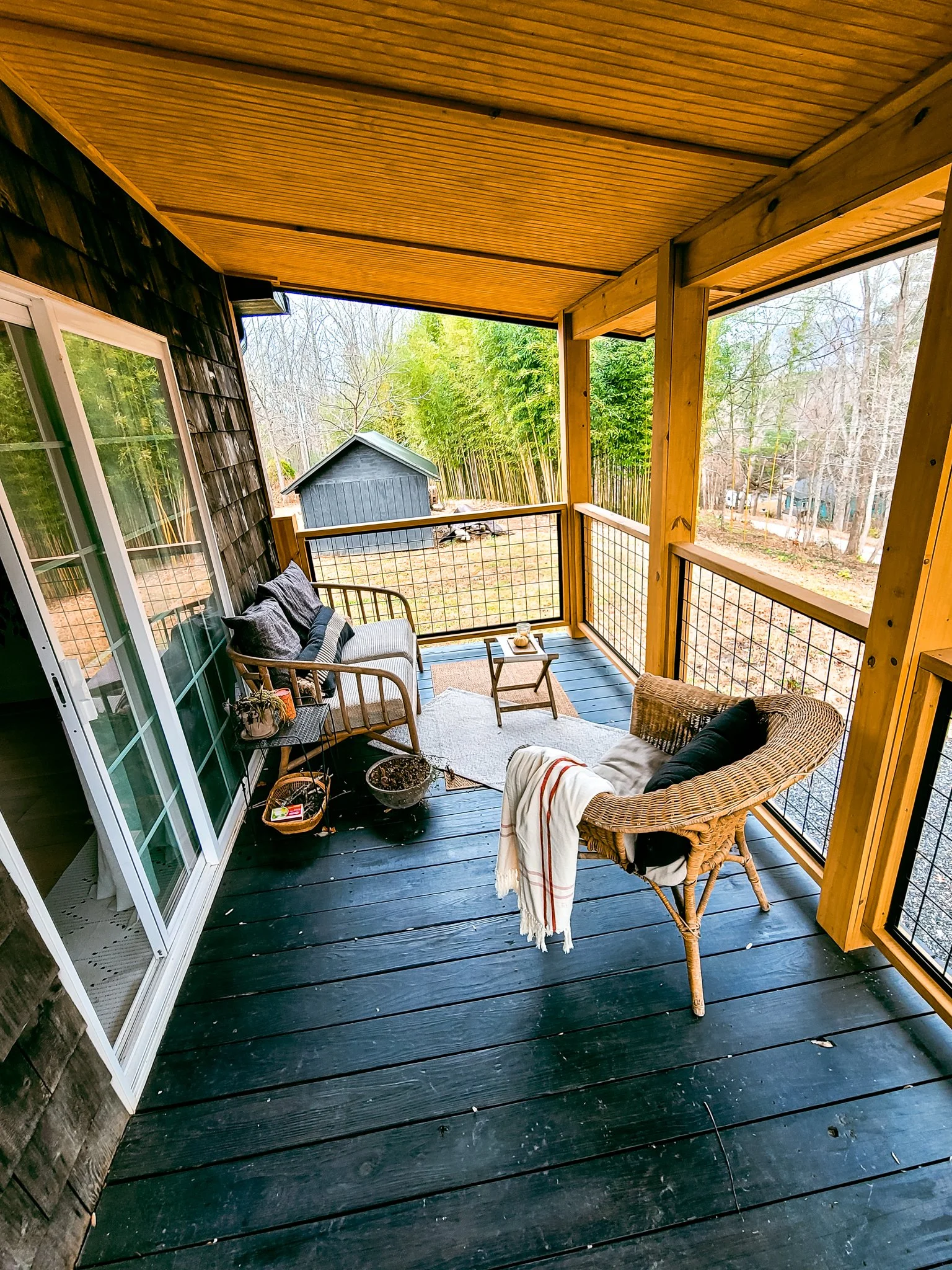 A cozy screened-in porch with black wooden floorboards, wicker and wooden outdoor furniture, and a view of a backyard with trees and a small shed.