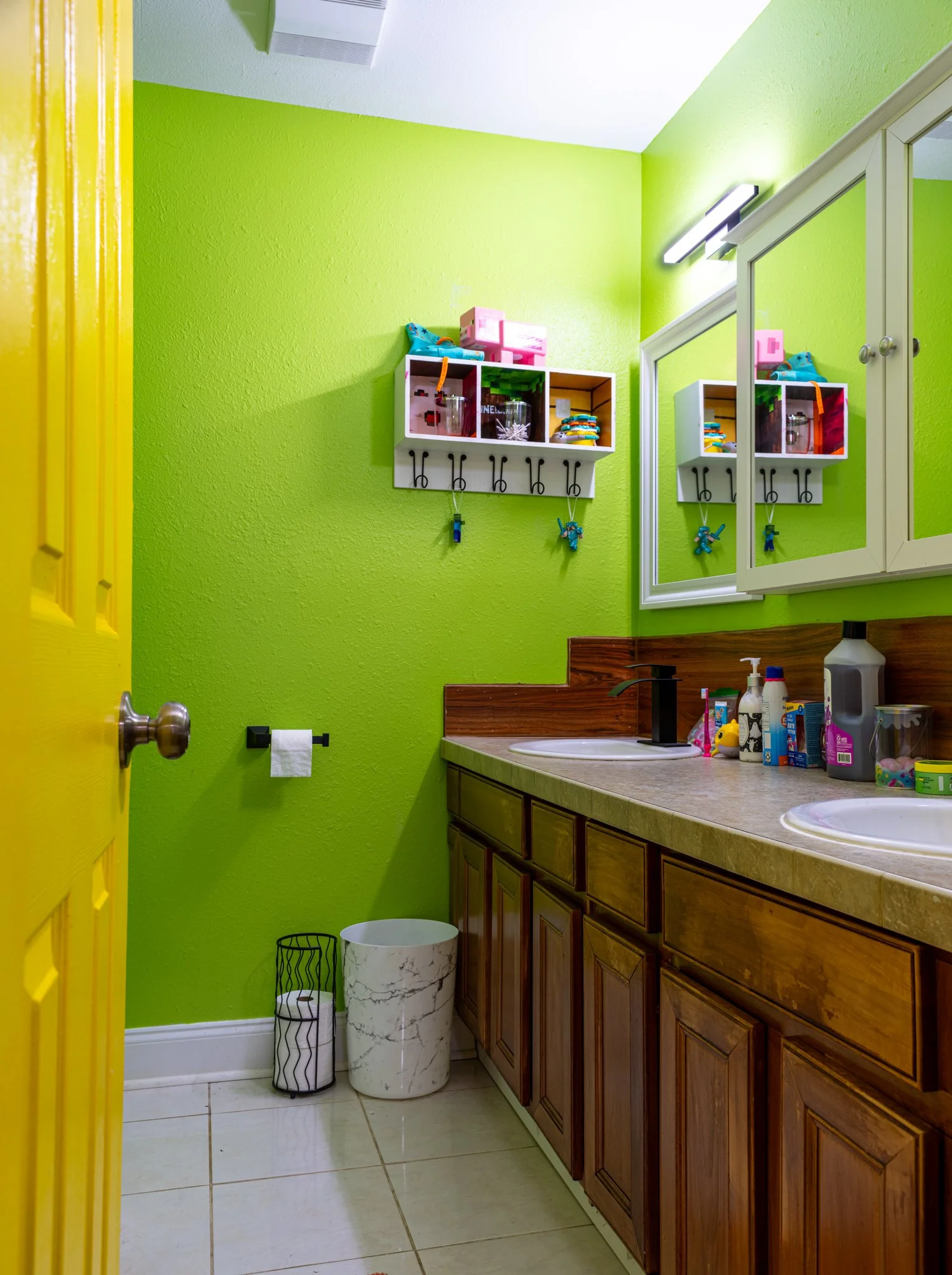 A brightly colored bathroom with lime green walls, a wooden vanity with two sinks, a large mirror, and a small cabinet. Decor includes storage boxes, toys, and toiletries, with a yellow door partially open.