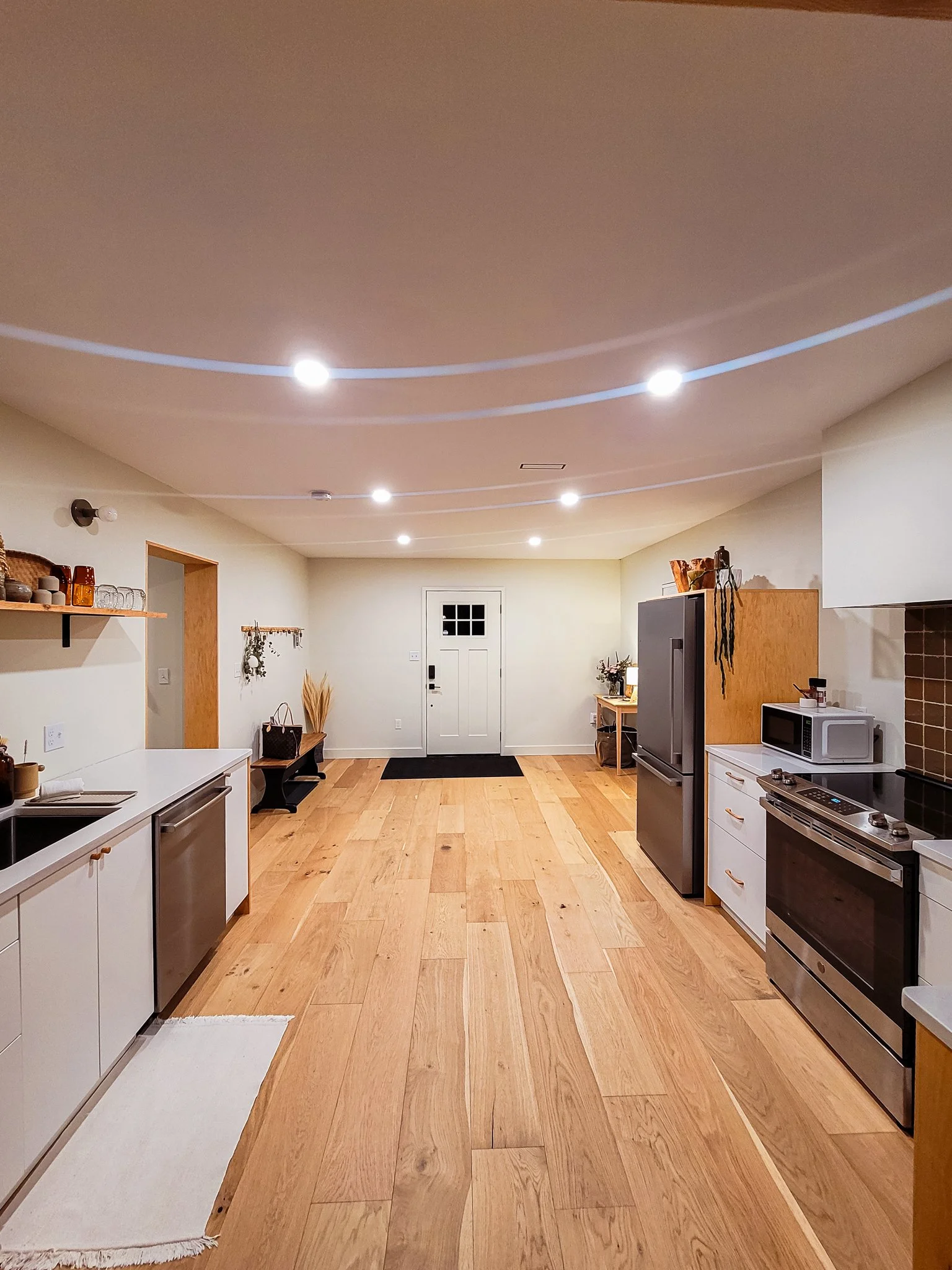 A spacious kitchen with a wooden floor, white cabinets, stainless steel appliances, and modern ceiling lighting. There is an entrance door at the far end with a black doormat in front of it.