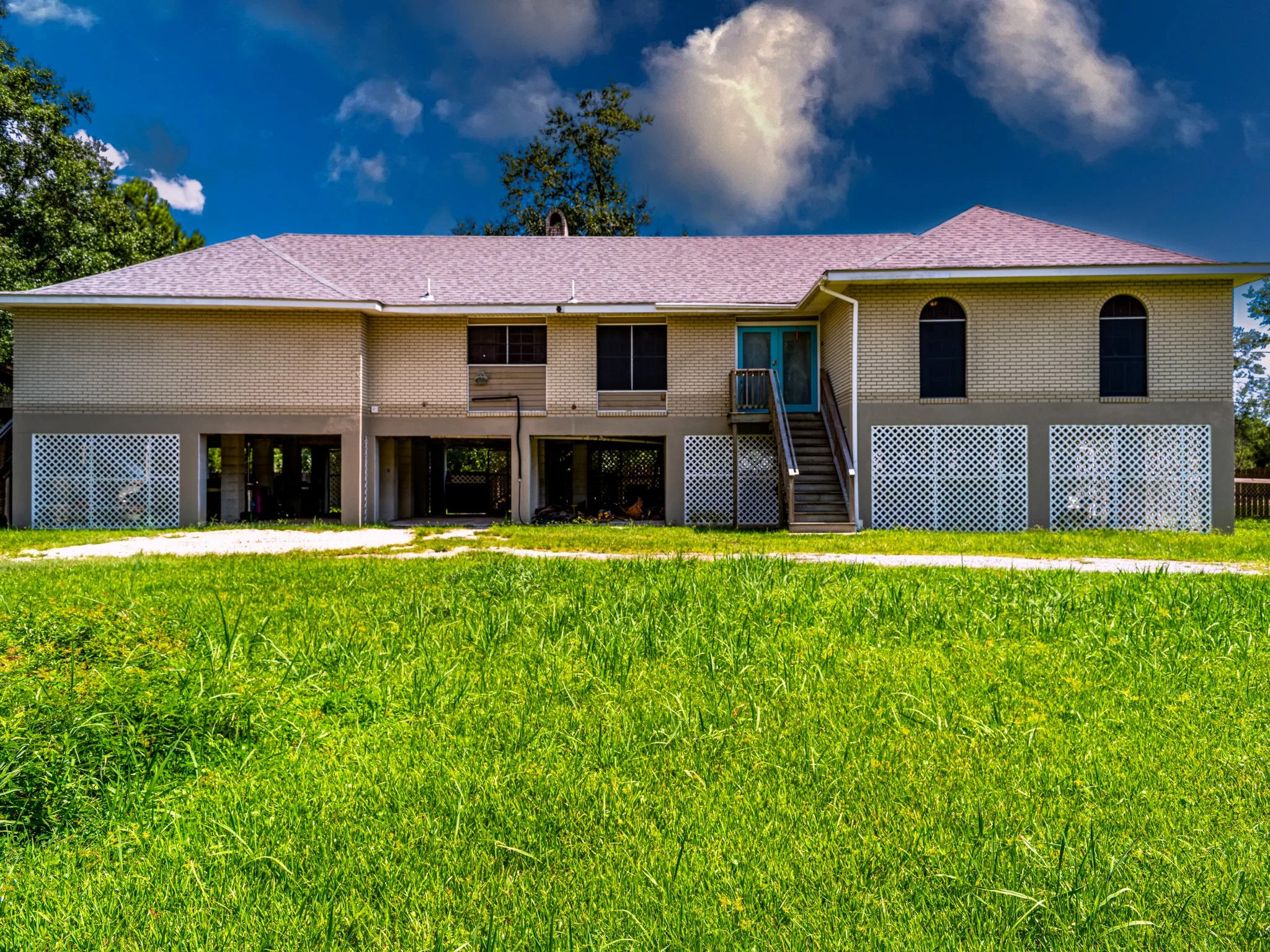 Back view of a two-story house with a pink roof, yellow brick exterior, and white lattice skirting, with a staircase leading to a screened door, under a partly cloudy sky.
