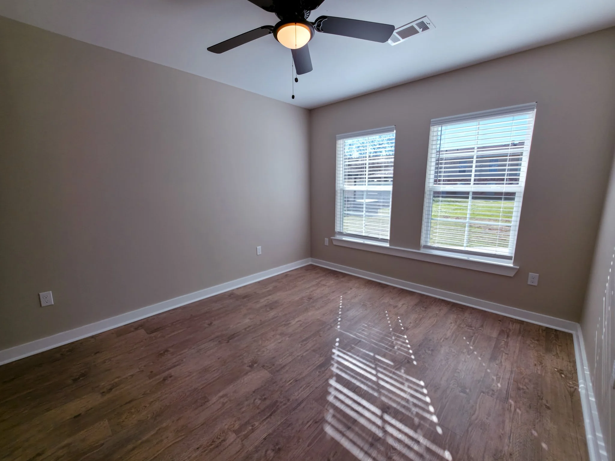 Empty room with beige walls, hardwood flooring, two windows with white blinds, a ceiling fan with a light, and sunlight streaming through the windows, creating shadows on the floor.