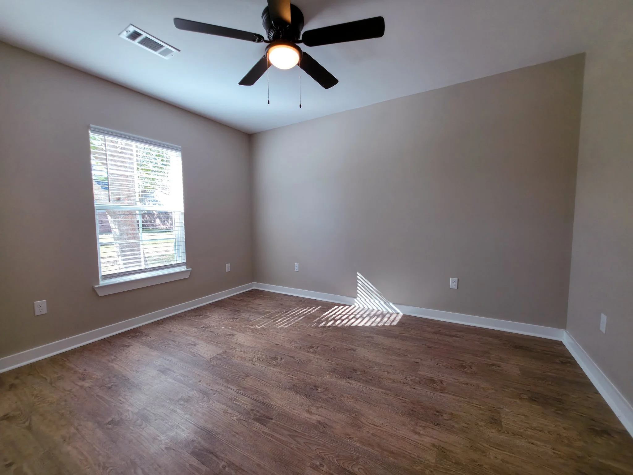 Empty room with hardwood floor, beige walls, white baseboards, a window with blinds, and a ceiling fan with a light