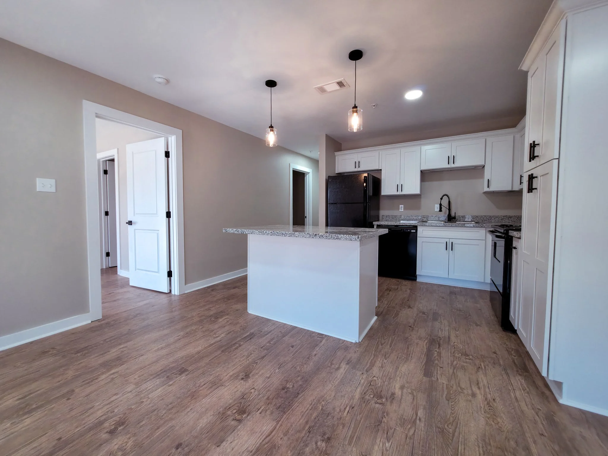 Empty kitchen with white cabinets, black appliances, granite countertops, wooden floor, and two pendant lights hanging from ceiling.