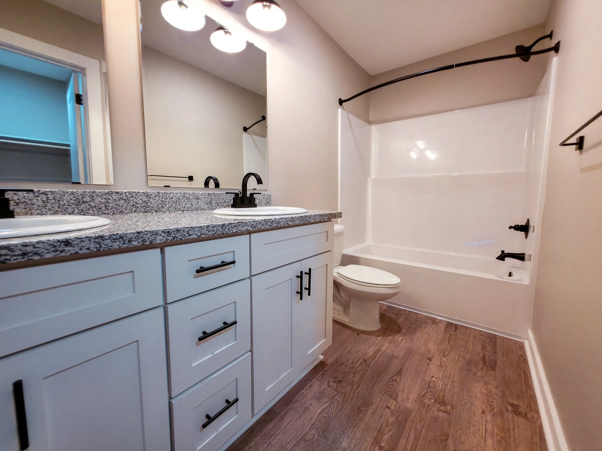 Bathroom with double vanity, granite countertop, black faucets, large mirror, toilet, bathtub with shower, dark curtain rod, and wood-look flooring.