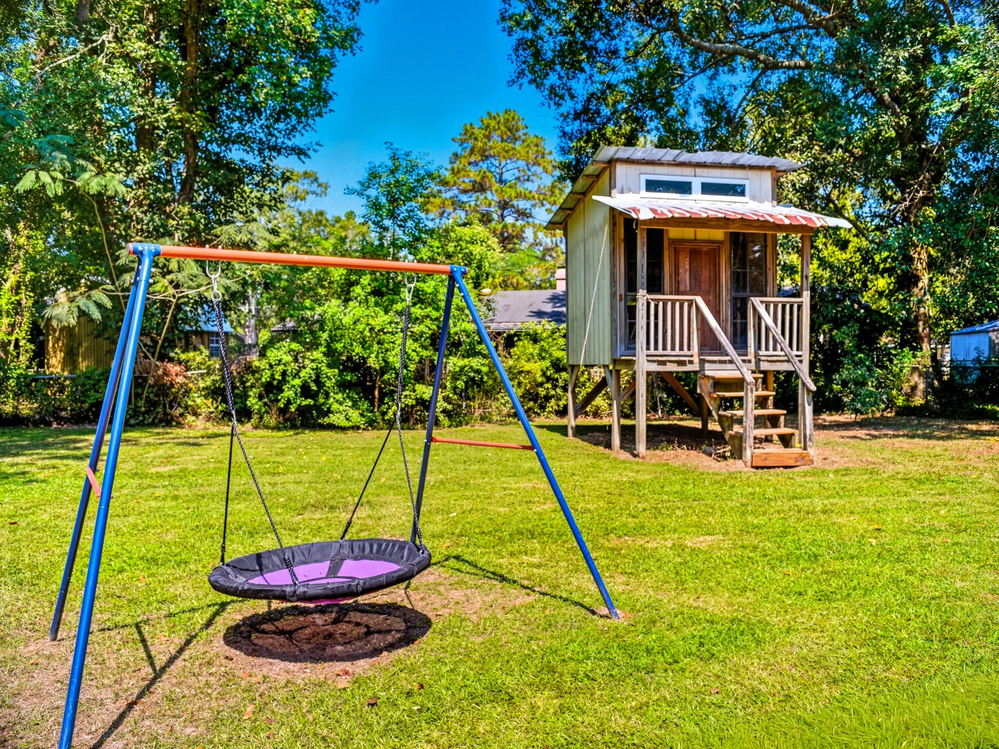 Colorful backyard with a playground swing set and a small wooden playhouse surrounded by green trees.