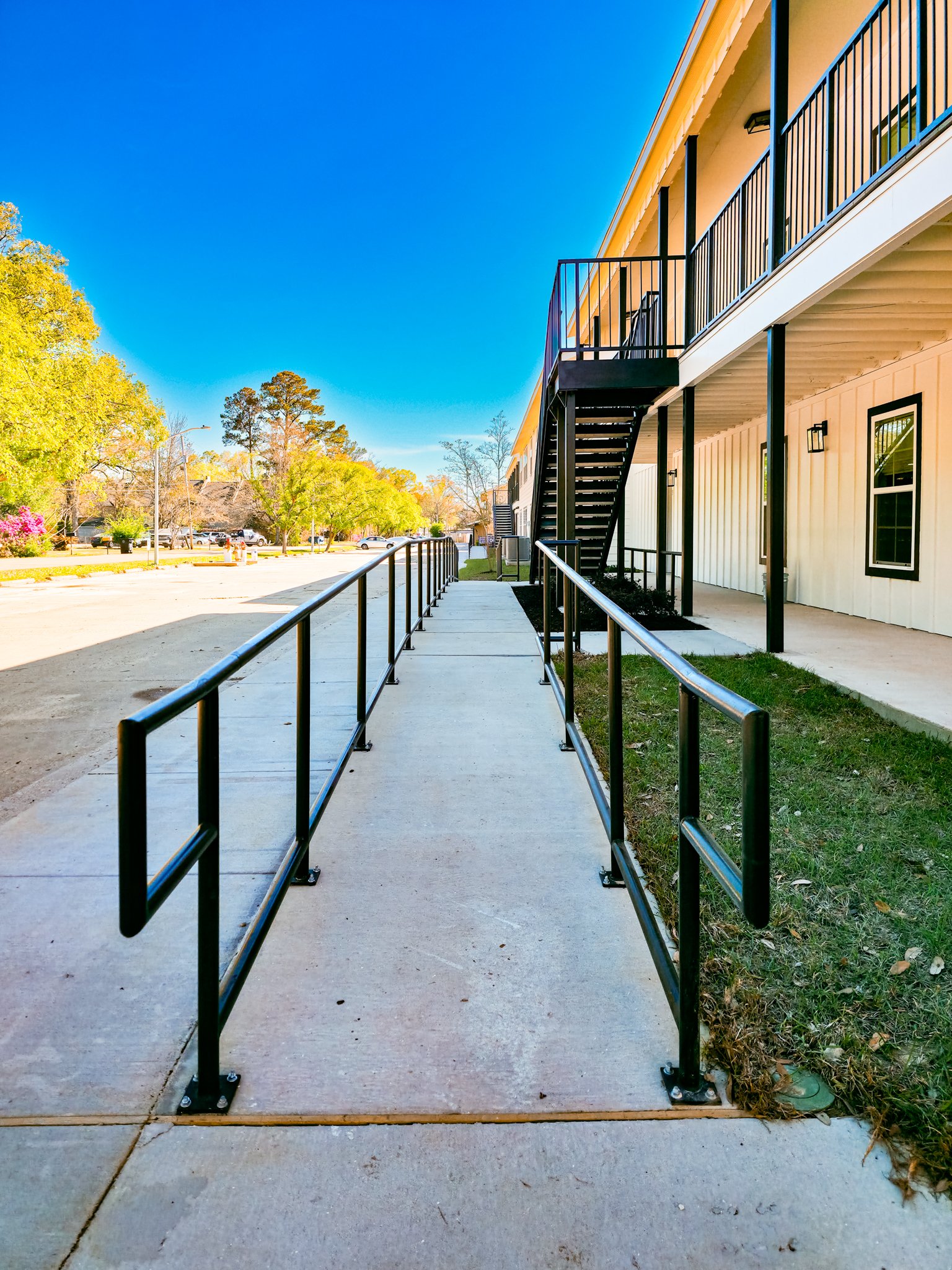 Exterior view of a residential building with a wheelchair accessible ramp leading to the upper floors, surrounded by trees and a parking lot under a clear blue sky.