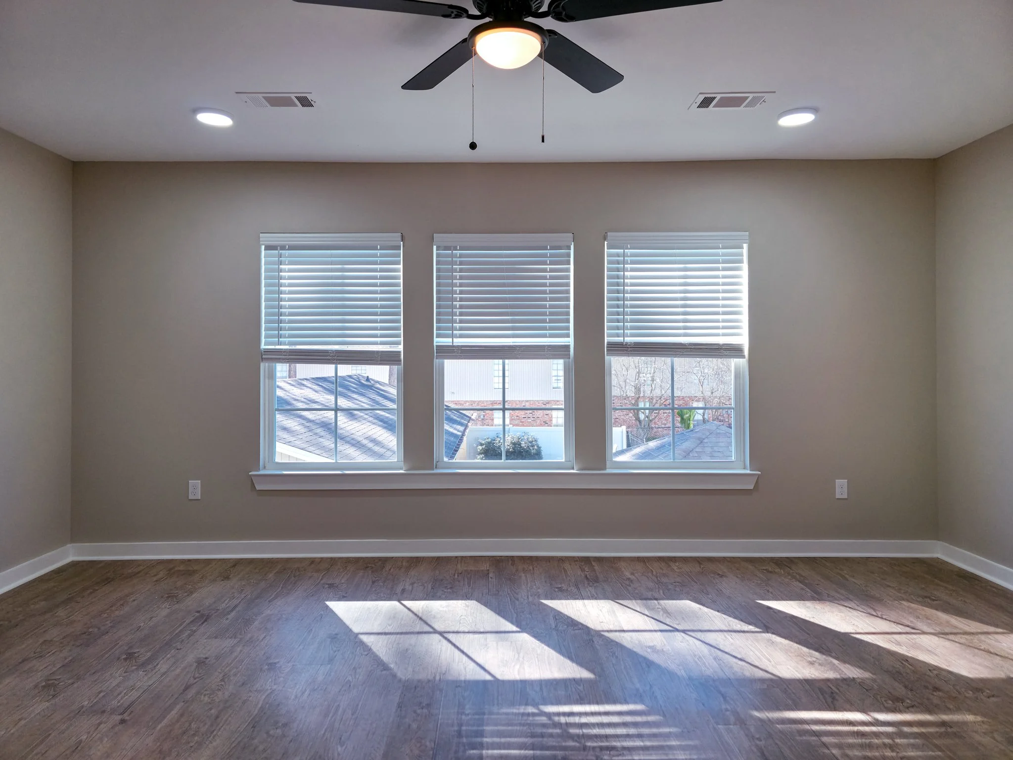 Empty room with three windows, blinds partially open, sunlight casting shadows on wooden floor, ceiling fan, and recessed lights