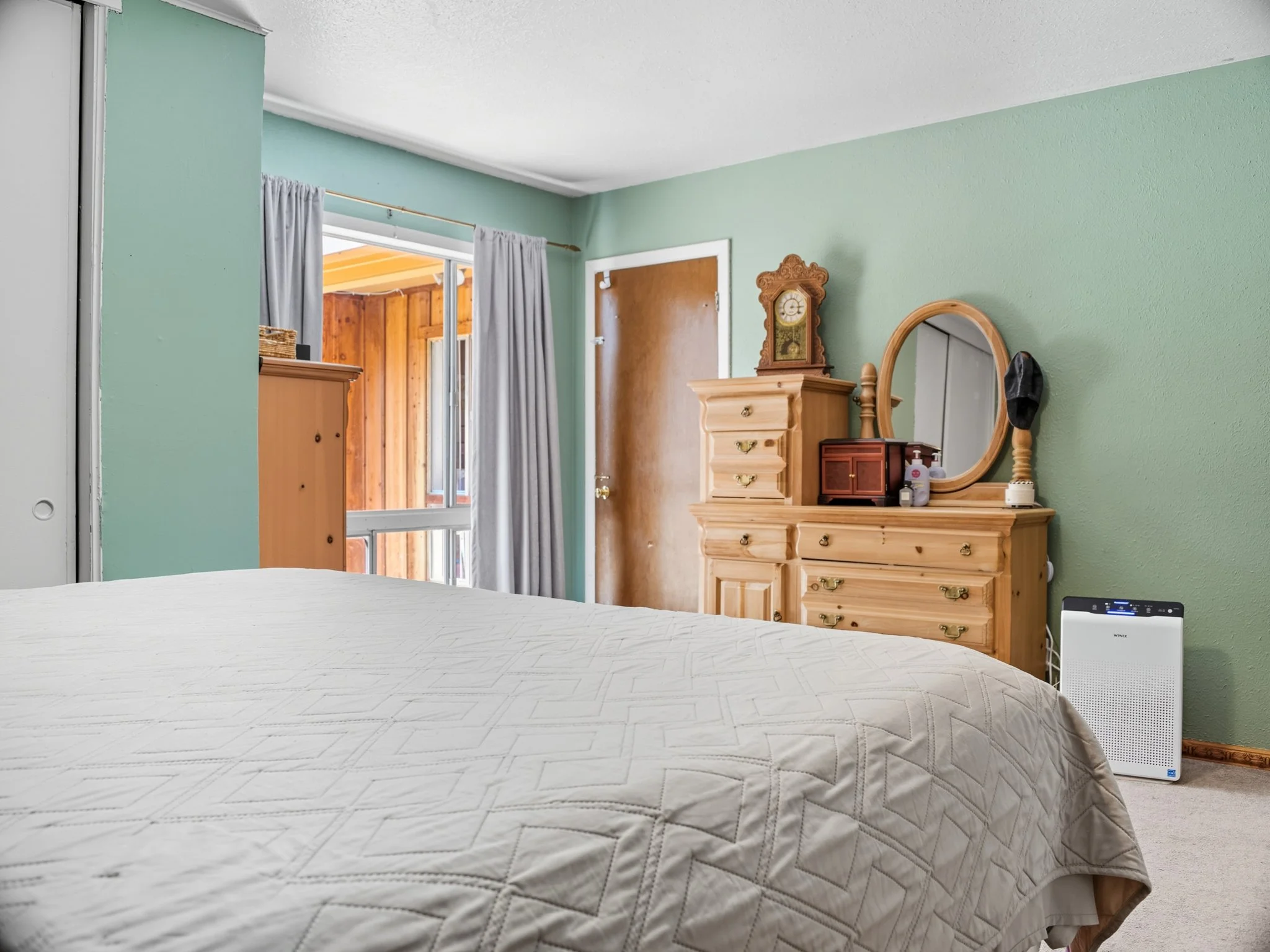 A bedroom with green walls, wooden furniture, a bed with a white quilt, a dresser with a mirror, a clock on top, clothes on the dresser, a wooden door, a window with curtains, and an air purifier.