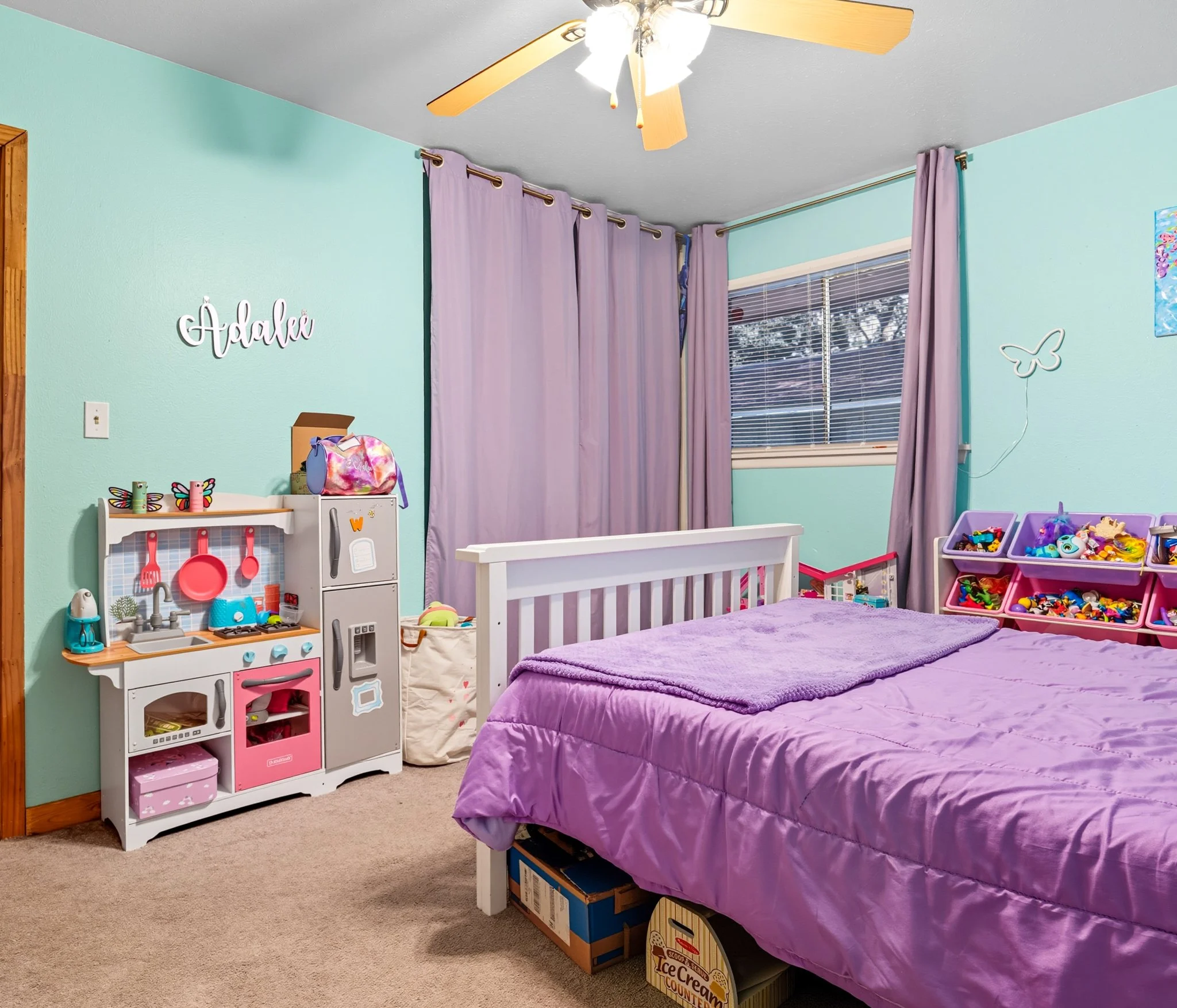A children's bedroom with light turquoise walls, purple curtains, and a purple bedspread. The room has toys, a toy kitchen, and storage bins filled with toys. A ceiling fan is above, and there's a window with blinds letting in natural light.