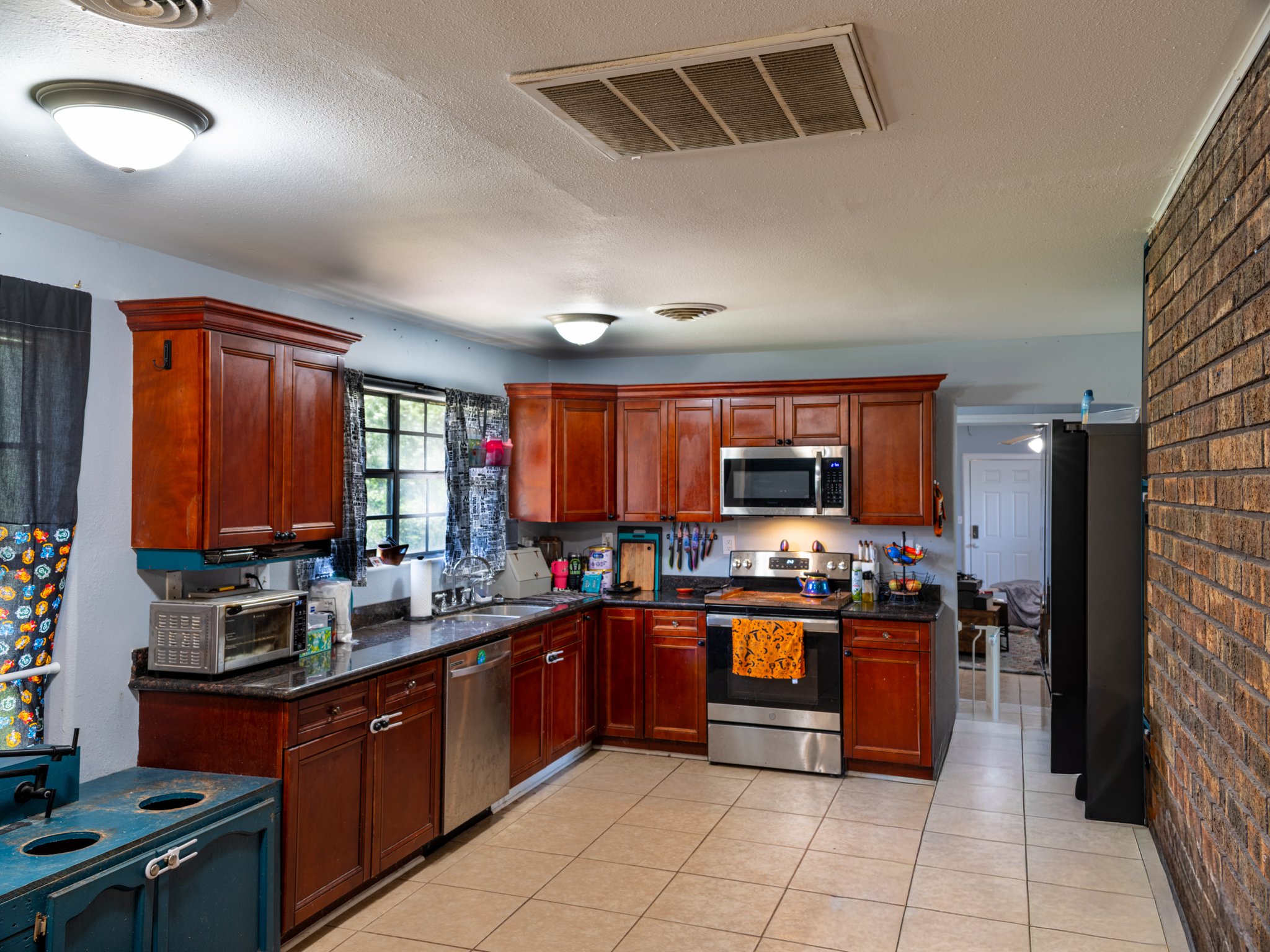 Kitchen with wooden cabinets, stainless steel appliances, black countertop, tiled floor, brick wall, window with curtains, microwave, toaster, and various kitchen items.