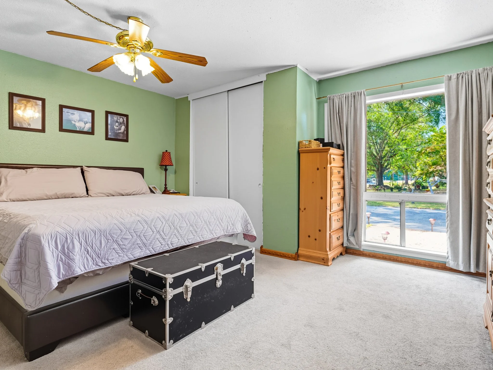 Bedroom with green walls, a large bed with beige pillows and a white quilt, a black trunk at the foot of the bed, a wooden dresser, a small wood nightstand with a red lamp, framed photos on the wall, a ceiling fan with wooden blades, beige curtains, 