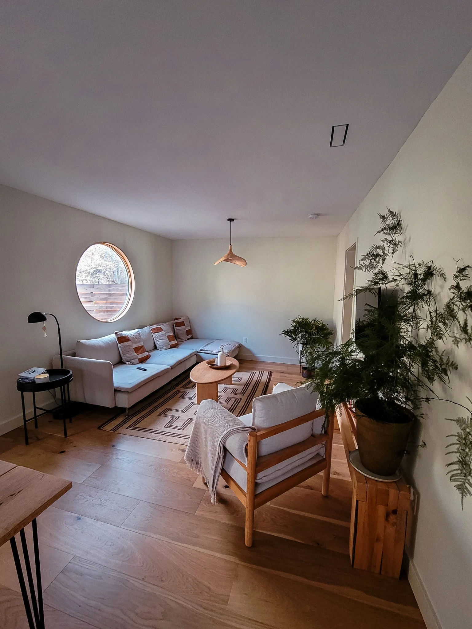 Living room with a white sectional sofa, wooden coffee table, and armchair, decorated with neutral tones, a round window, and numerous indoor plants.