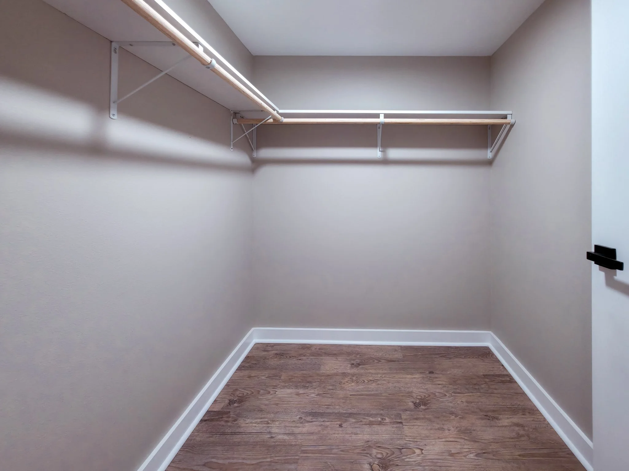 Empty walk-in closet with beige walls, wooden flooring, and white shelving rods on both sides.