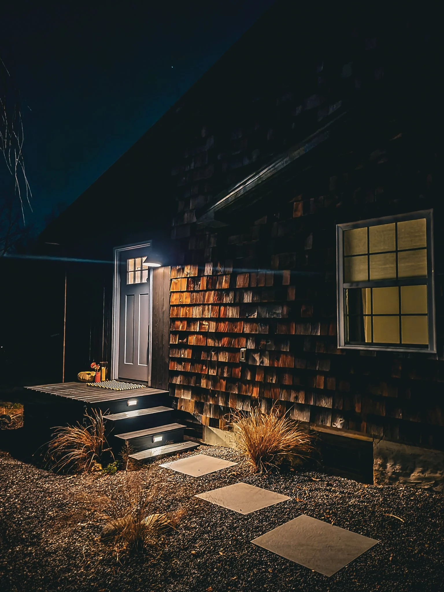 Nighttime exterior view of a house with a dark door, illuminated by warm outdoor lighting, wooden shingle siding, window with curtains, stone pathway, and plants in the yard.
