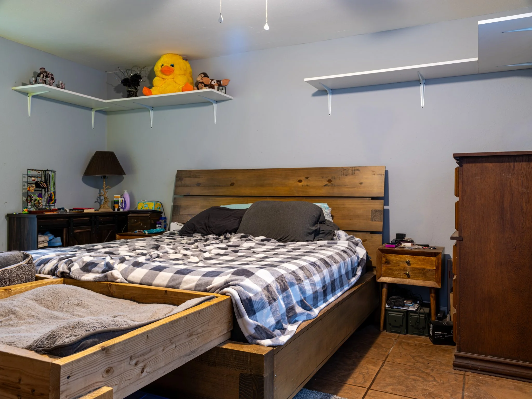 A bedroom with a wooden bed, a checkered blanket, and pillows. There are two shelves with stuffed animals and decorations, a nightstand with items and a dresser in the corner. The room has tiled flooring.