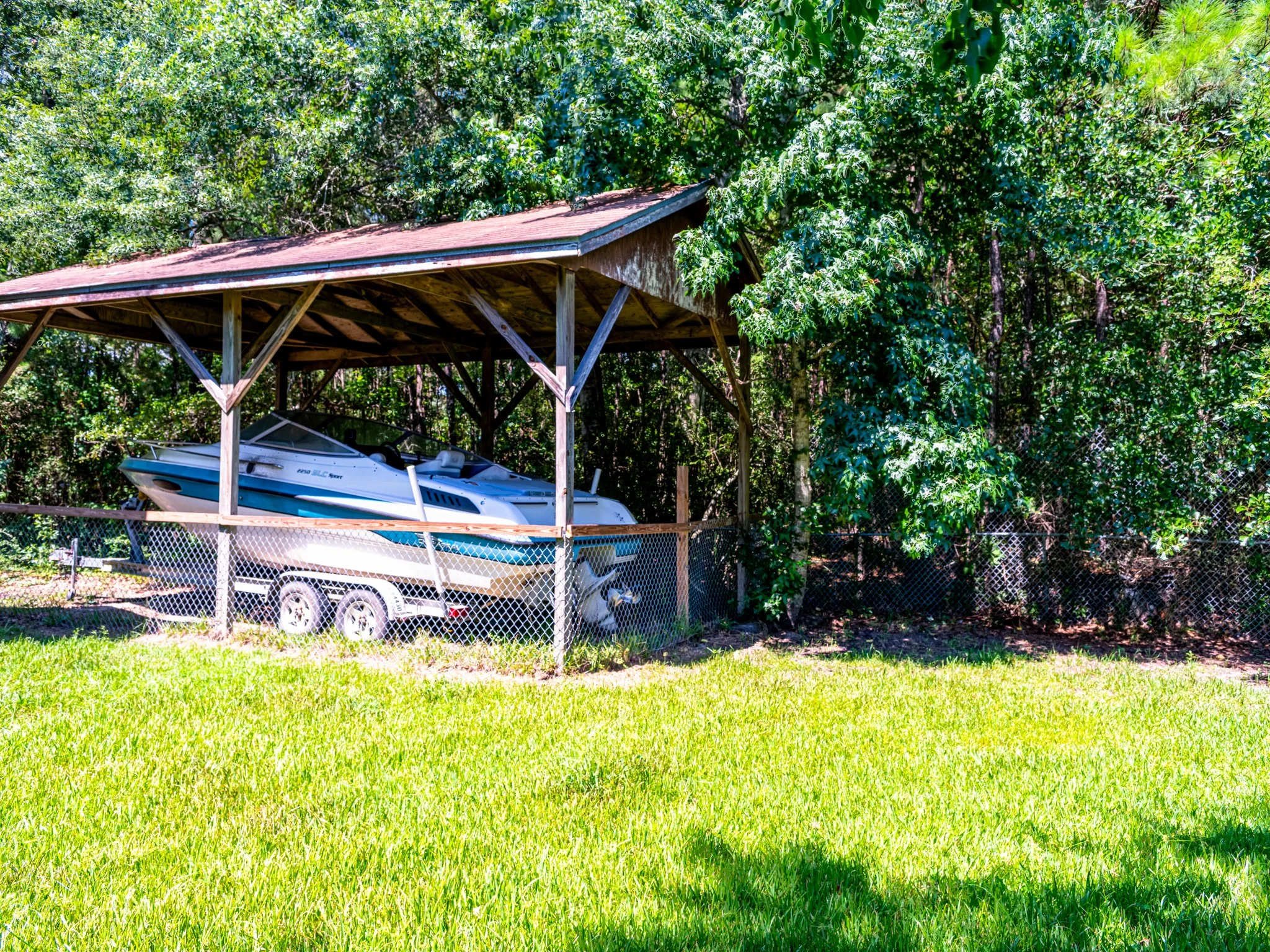 Boat stored under a wooden shelter on a trailer, next to a grassy yard with trees and a chain-link fence.