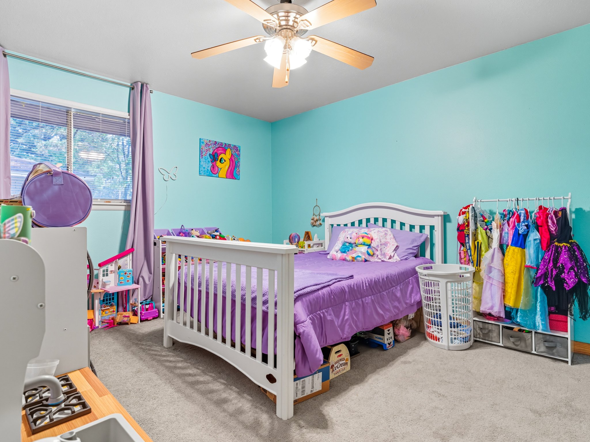 A child's bedroom with a white bed, purple bedding, and colorful toys. Lilac curtains frame a window with a view of trees. A wall decoration features a cartoon pony. A clothing rack holds princess costumes. A laundry basket is beside the bed. Fluffy 