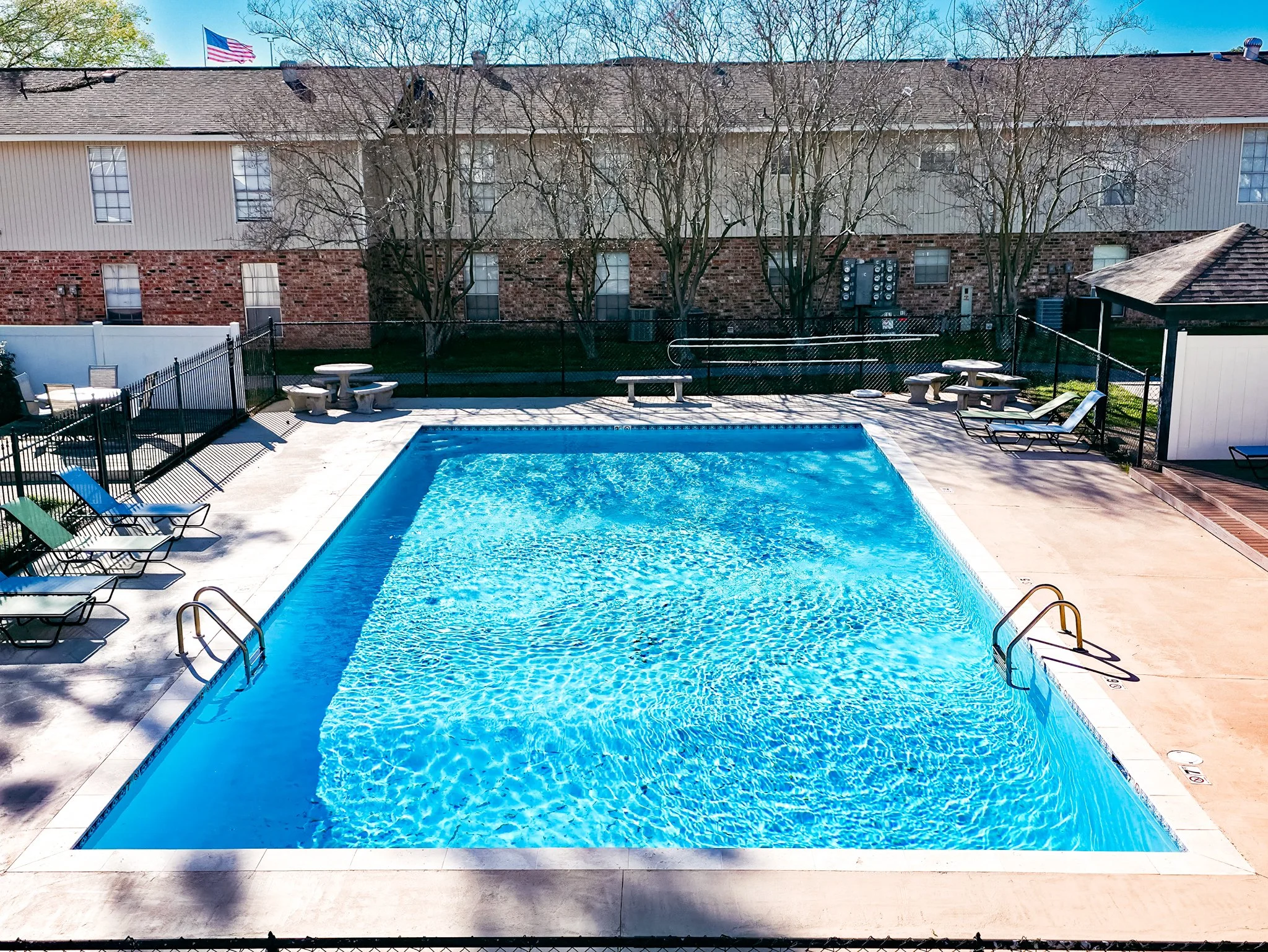 Empty outdoor swimming pool with clear blue water, surrounded by concrete deck and lounge chairs, with leafless trees and a brick apartment building in the background.