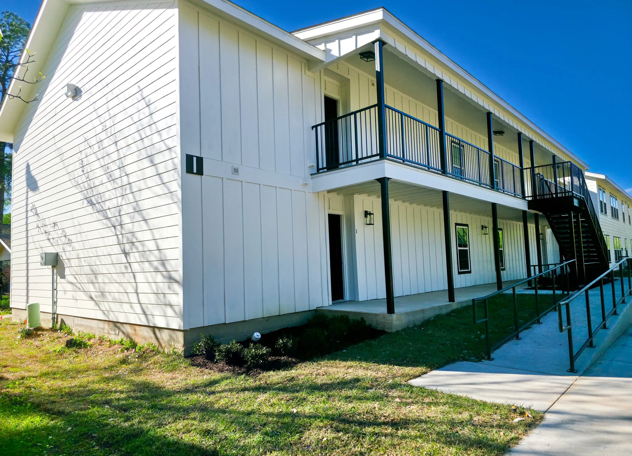 Exterior view of a modern, white two-story apartment building with black railings and stairs, under a clear blue sky.