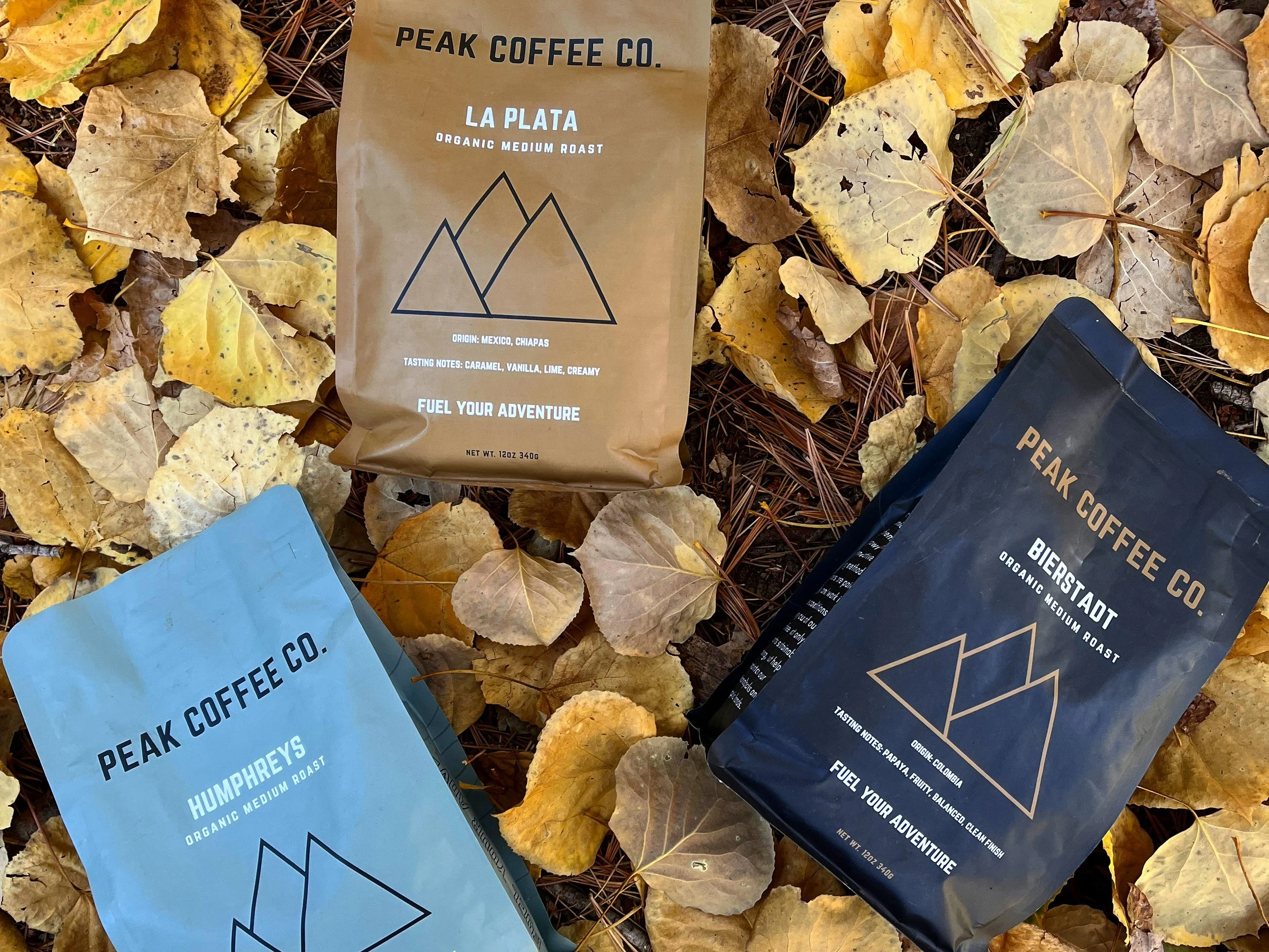 Three bags of Peak Coffee Co. coffee on autumn leaves. The bags are labeled 'La Plata,' 'Humphrey's,' and 'Bierstadt,' each indicating their organic medium roast with different tasting notes. The La Plata is from Mexico, Chiapas with caramel, vanilla, lime, and creamy notes. Humphrey's is from Colombia with Padoca, fruity, balanced, and clean finish. Bierstadt is from Colombia with papaya, fruity, balanced, and clean finish.