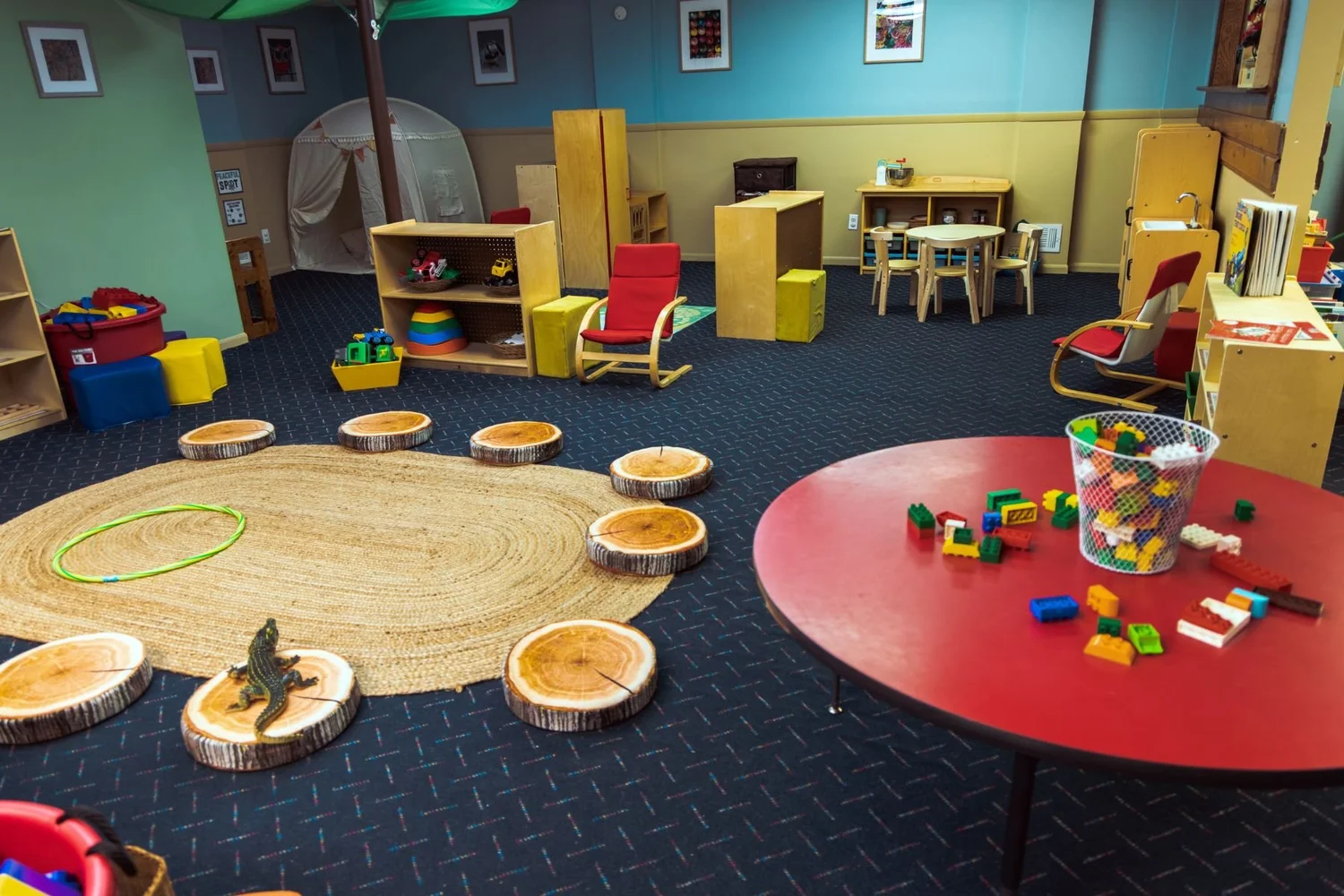 Early preschool room with a woven rug, tree-stump seats, and a red table