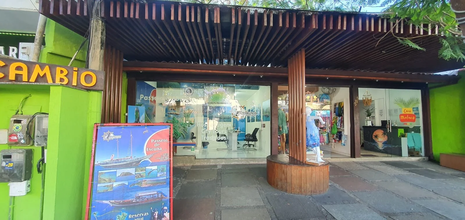 Fachada de loja de turismo com vidro na frente, decoração de madeira e um manequim com roupa azul e chapéu. Ao lado, sinal de câmbio de moeda e um painel de publicidade de passeios de escuna e reservas.